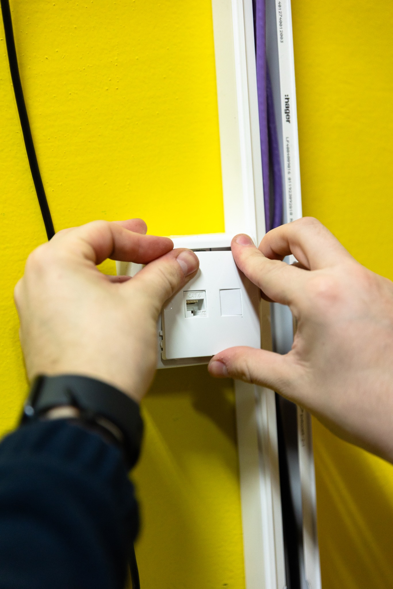 Montaż gniazda sieciowego RJ45 na żółtej ścianie, widoczne ręce instalatora, listwa przypodłogowa z kablem. Instalacja sieci LAN w biurze lub domu.