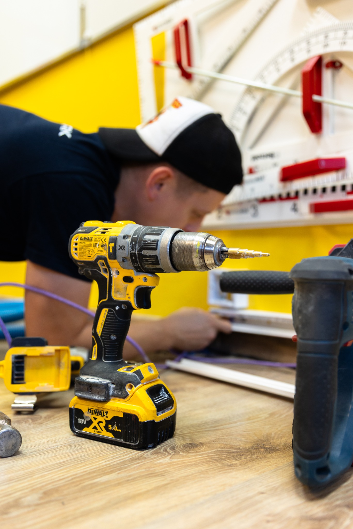 Elektryk w trakcie pracy, wiertarka DeWalt na pierwszym planie, żółta ściana i narzędzia w tle. Precyzyjne wiercenie otworów, instalacja elektryczna w trakcie realizacji.