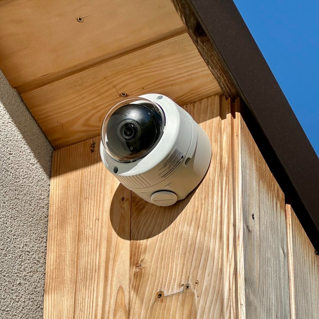 Kamera kopułkowa monitoringu, zamontowana na drewnianej elewacji budynku, widoczna z dołu pod kątem, z fragmentem nieba w tle. Jasne oświetlenie.