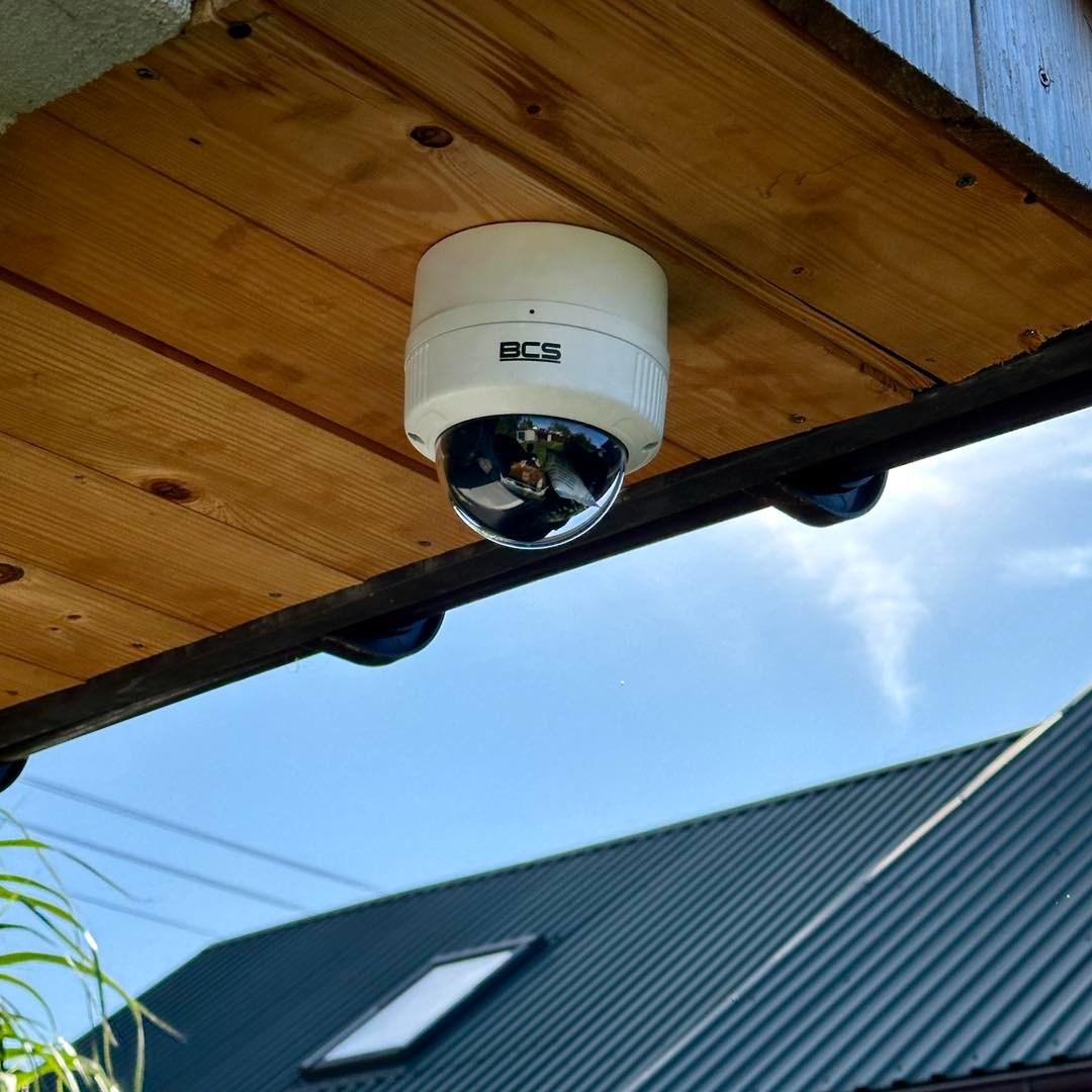 Kamera kopułkowa BCS zamontowana pod drewnianym zadaszeniem, widok z dołu na tle błękitnego nieba i dachu domu. Instalacja monitoringu wizyjnego.