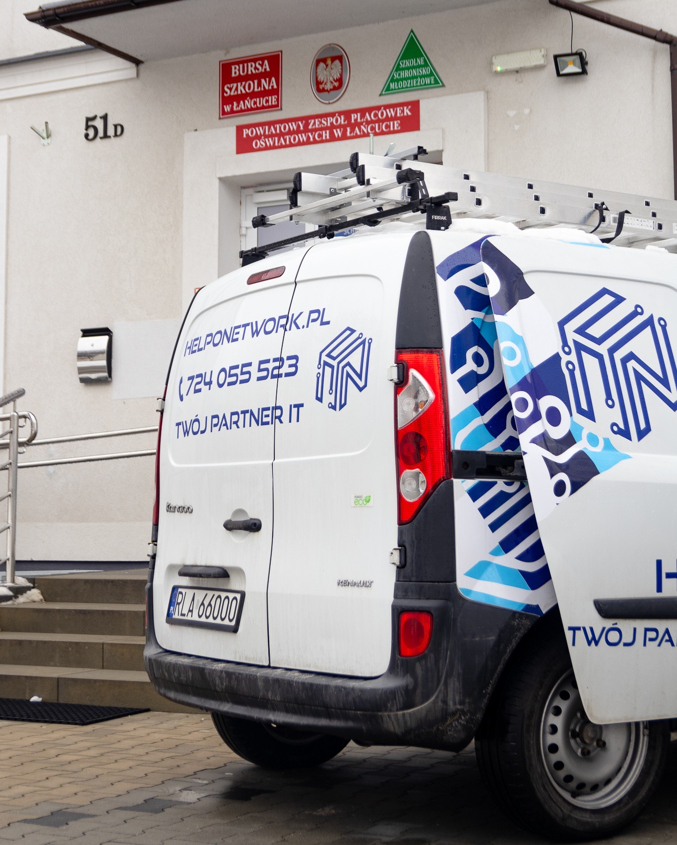 Biały samochód dostawczy z logo i danymi kontaktowymi firmy IT na tle budynku z tablicami informacyjnymi. Na dachu drabina. Widoczny numer telefonu i adres strony internetowej.