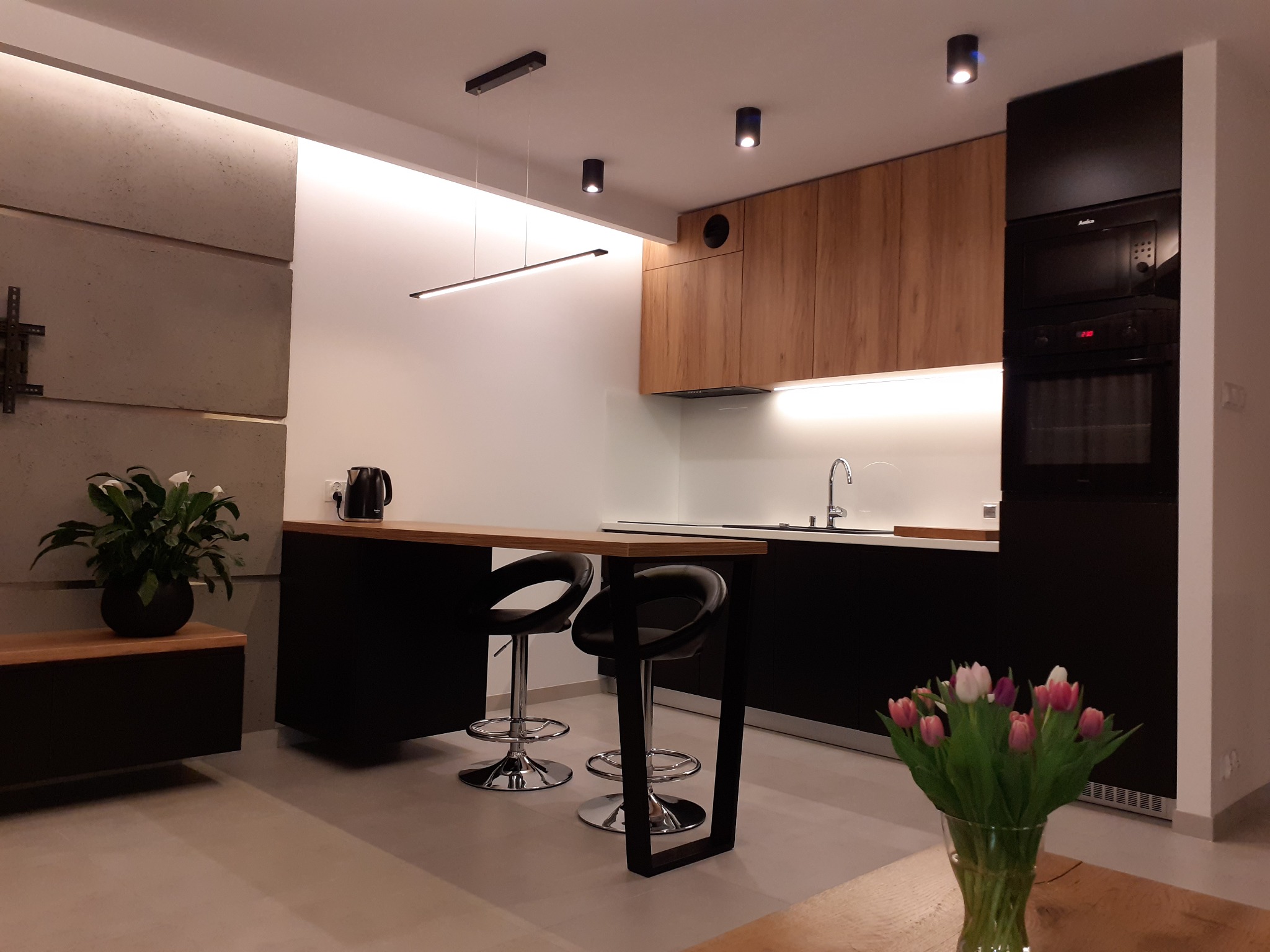 Nowoczesna kuchnia z drewnianymi szafkami, czarnymi elementami i wyspą z hokerami. Minimalistyczne oświetlenie i dekoracje roślinne dopełniają wnętrze.