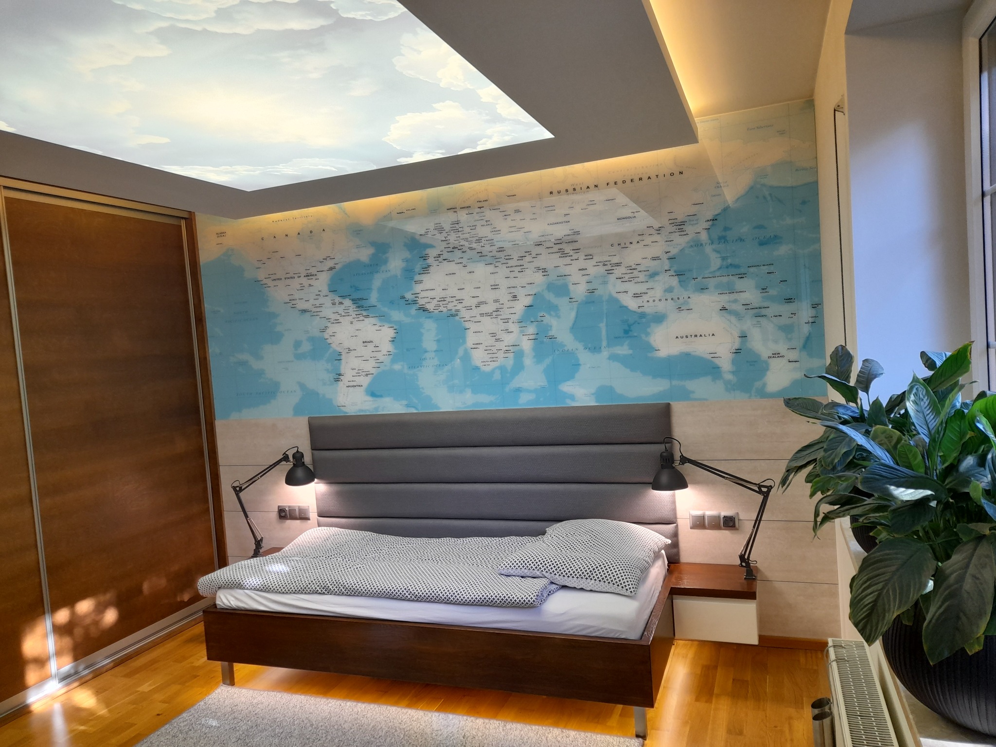 Sypialnia z łóżkiem, mapa świata na ścianie i podświetlanym sufitem z motywem chmur. Nowoczesny design wnętrza z drewnianą podłogą i dużą rośliną doniczkową.
