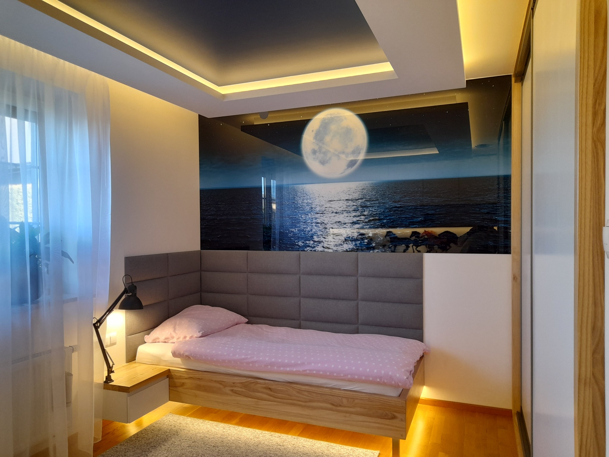 Nowoczesna sypialnia z łóżkiem, panelem tapicerowanym i fototapetą z motywem księżyca nad morzem. Oświetlenie LED w suficie i pod szafką nocną. Drewniana podłoga.