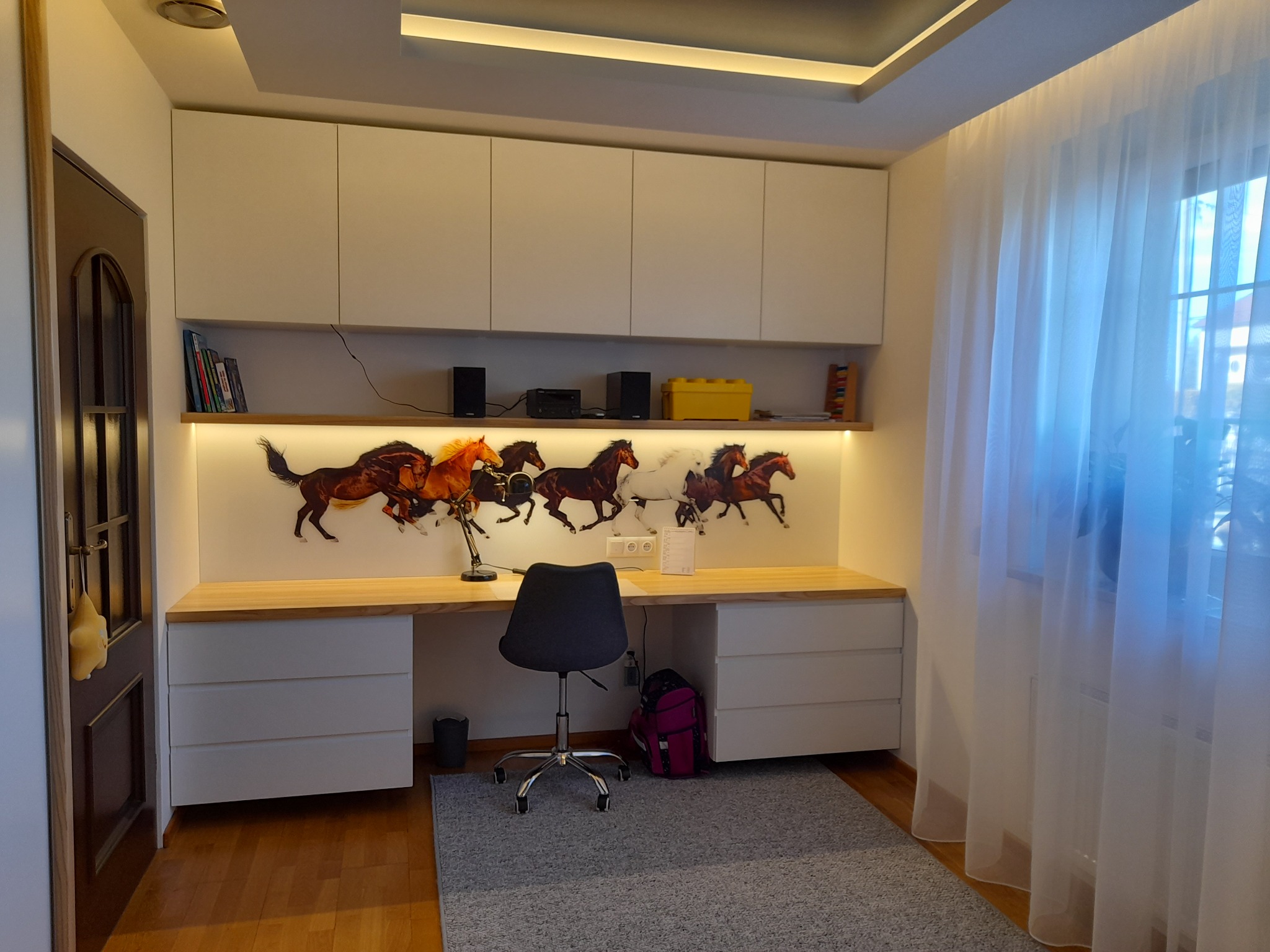 Białe szafki nad biurkiem z motywem koni, oświetlenie LED, szare krzesło i dywan. Nowoczesne wykończenie wnętrza pokoju z zasłonami przy oknie.