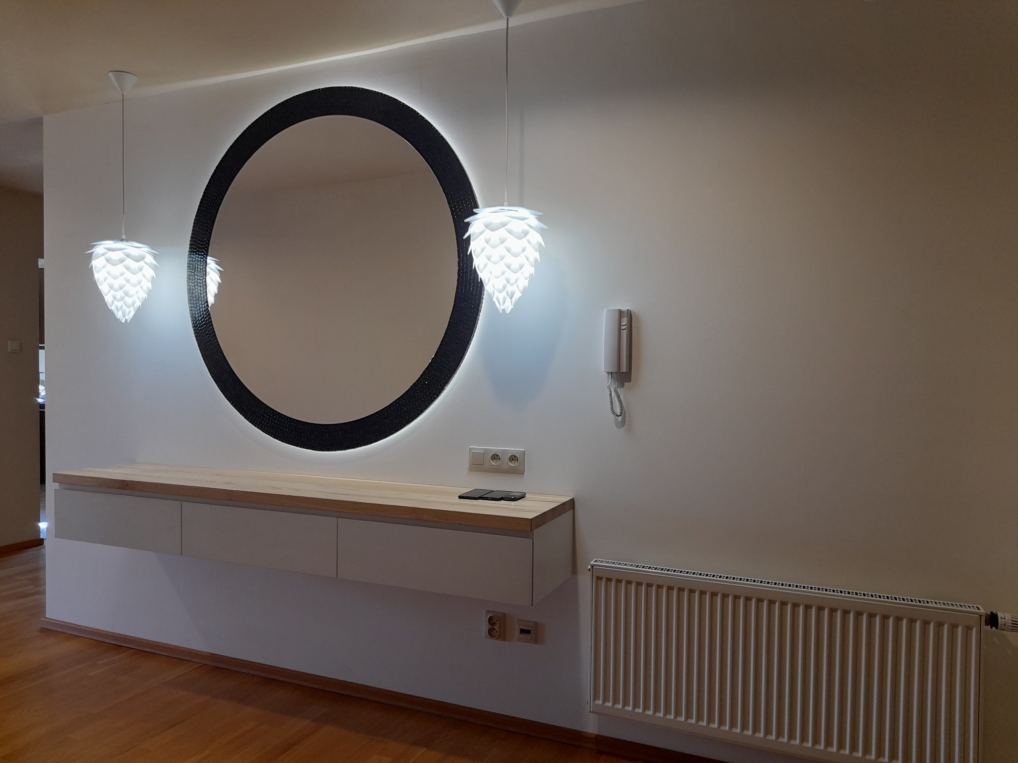 Nowoczesny przedpokój z okrągłym lustrem z podświetleniem LED, wiszącą szafką z drewnianym blatem i grzejnikiem. Dwie lampy w kształcie szyszki. Minimalistyczny design.
