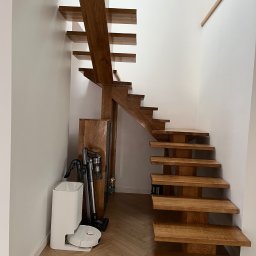 Pracownia stolarska Kamil pryk - Nowoczesne, drewniane schody ażurowe z dębowymi stopniami, wsparte na solidnej konstrukcji. W tle białe ściany i parkiet w jodełkę. Widoczny odkurzacz automatyczny.