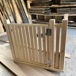 Pracownia stolarska Kamil pryk - Surowa, jasna, drewniana furtka z zawiasami, w warsztacie stolarskim. W tle deski i półki z drewnem. Widoczne niedoskonałości betonu.