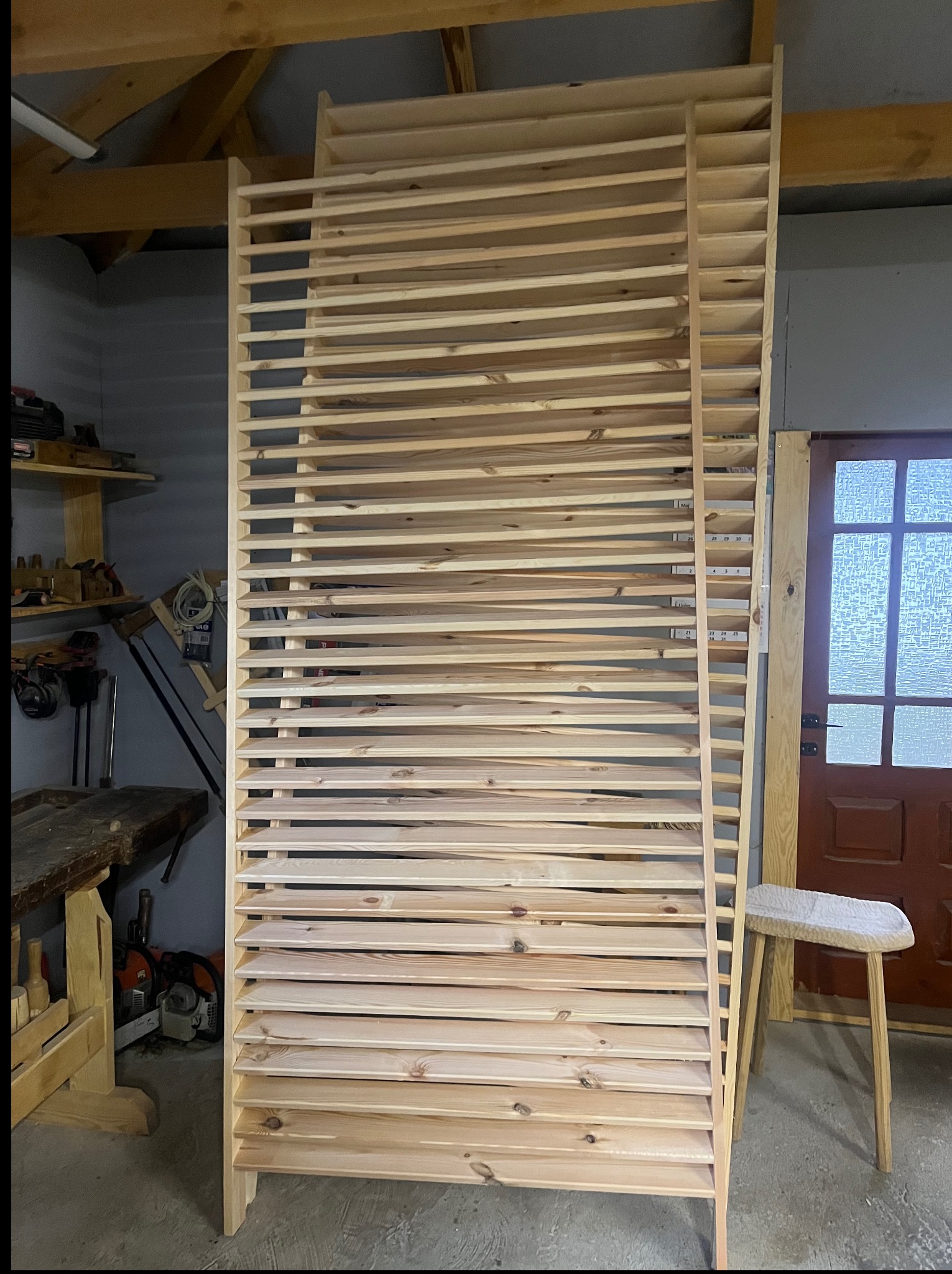 Surowa, drewniana konstrukcja z listew w warsztacie, prawdopodobnie element większej całości, z widocznymi narzędziami i stołkiem w tle. Jasne drewno, proste linie.