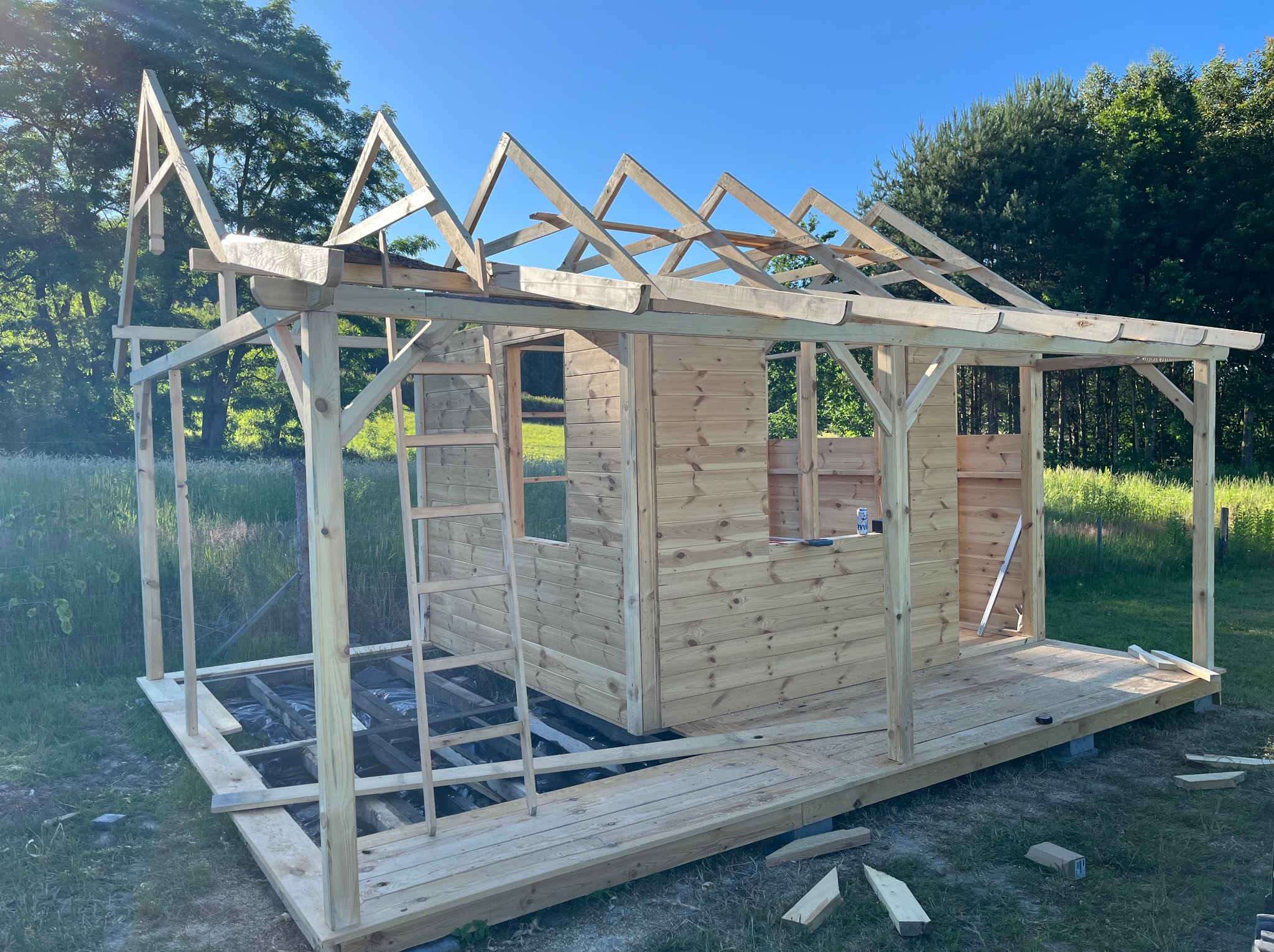 Wznoszenie drewnianej konstrukcji altany z widoczną konstrukcją dachu i ścian. Na pierwszym planie fragment tarasu. Prace w toku, widoczne narzędzia i drabina.