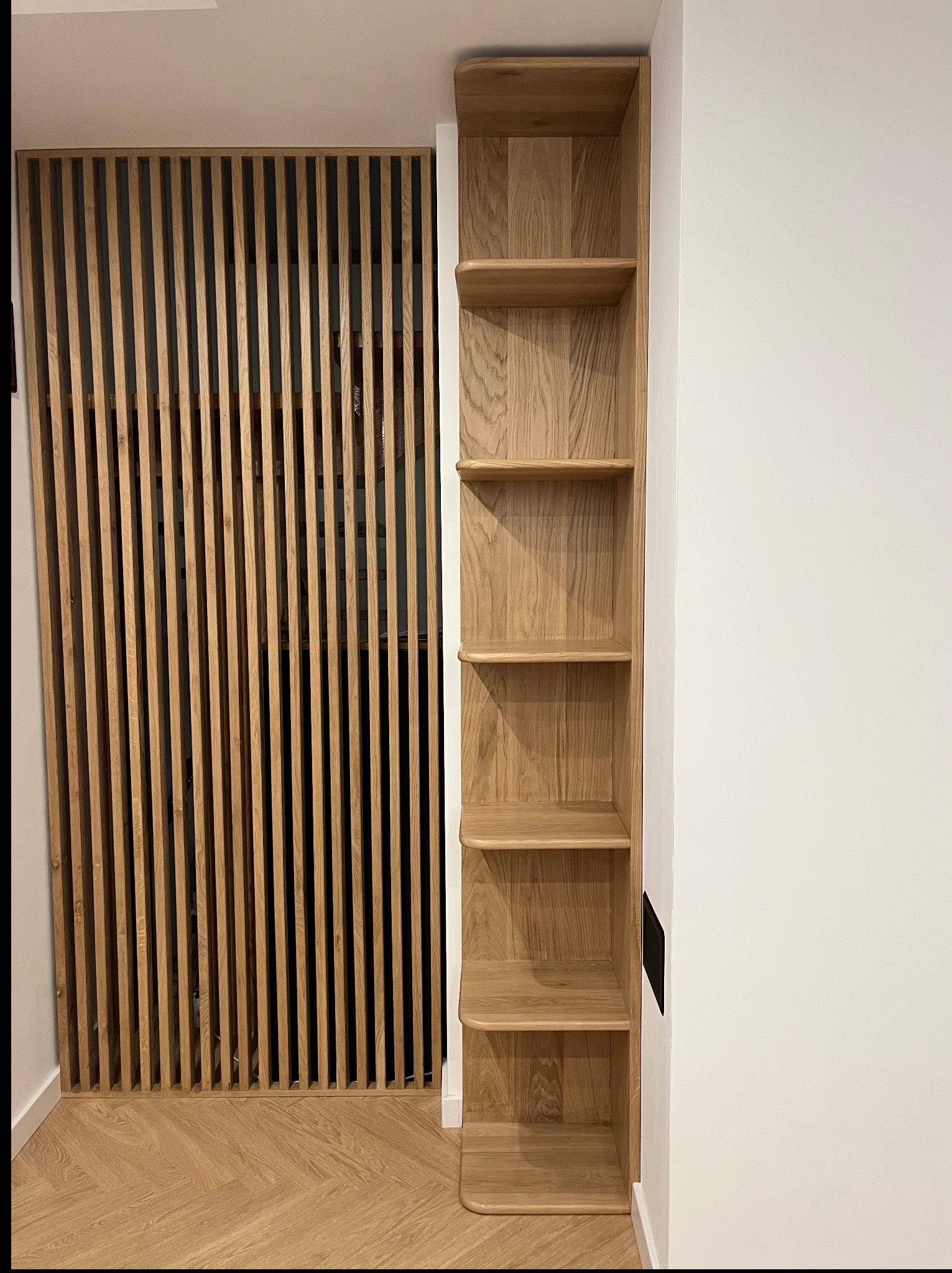 Drewniana zabudowa: ażurowa ścianka działowa z pionowych listew i wąski regał z półkami z litego drewna, dopasowane do parkietu w jodełkę. Minimalistyczny design.