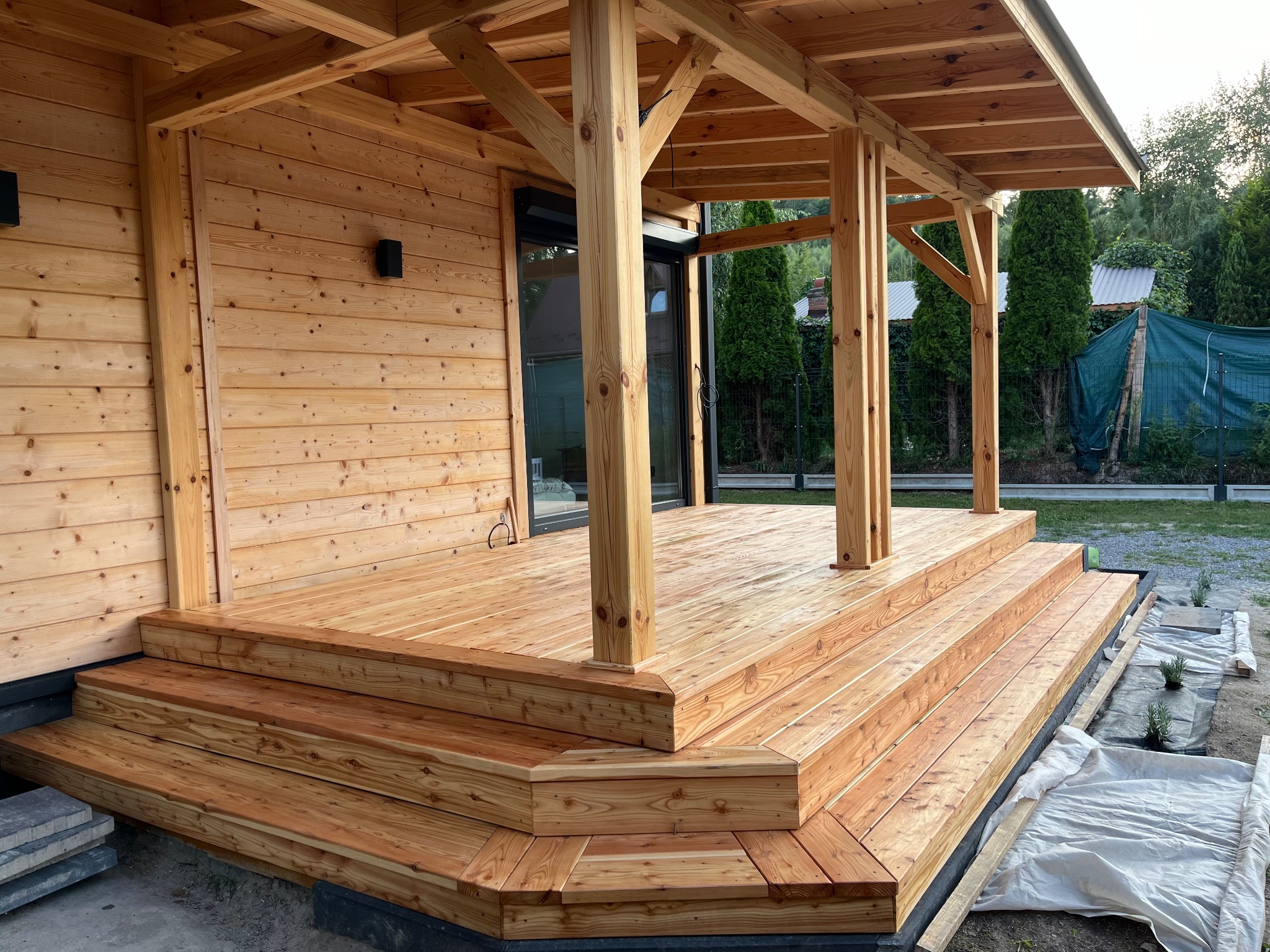 Jasny, drewniany taras z zadaszeniem, widok z dołu. Schody prowadzą na taras, w tle zieleń ogrodu. Widoczne detale konstrukcji i naturalne usłojenie drewna.