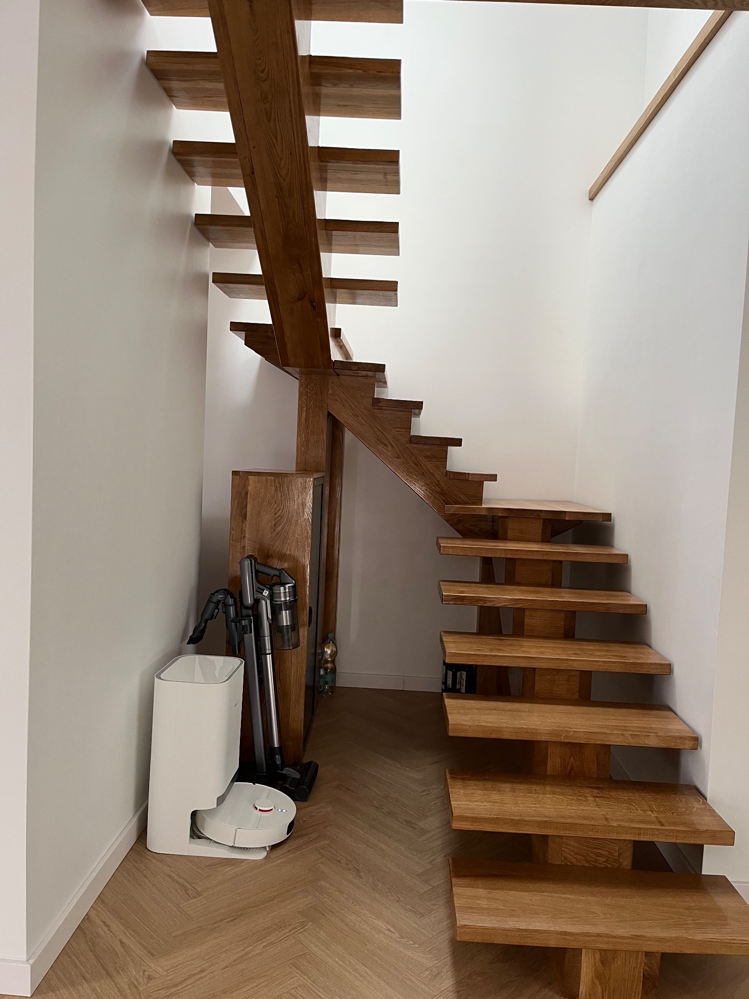 Nowoczesne, drewniane schody ażurowe z dębowymi stopniami, wsparte na solidnej konstrukcji. W tle białe ściany i parkiet w jodełkę. Widoczny odkurzacz automatyczny.