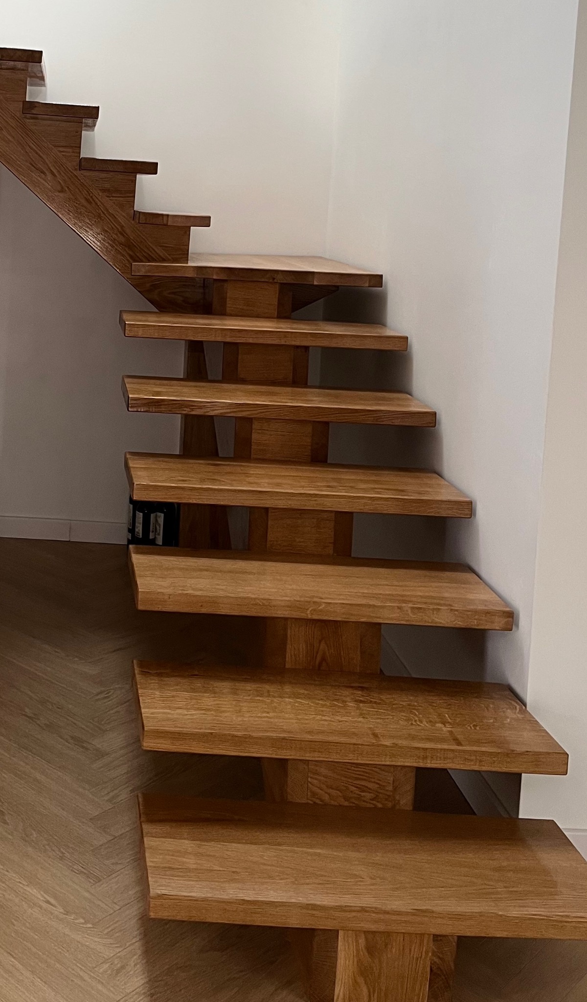 Nowoczesne, drewniane schody wspornikowe z dębowymi stopniami, zamontowane przy białej ścianie. Widoczny fragment parkietu. Minimalistyczny design.