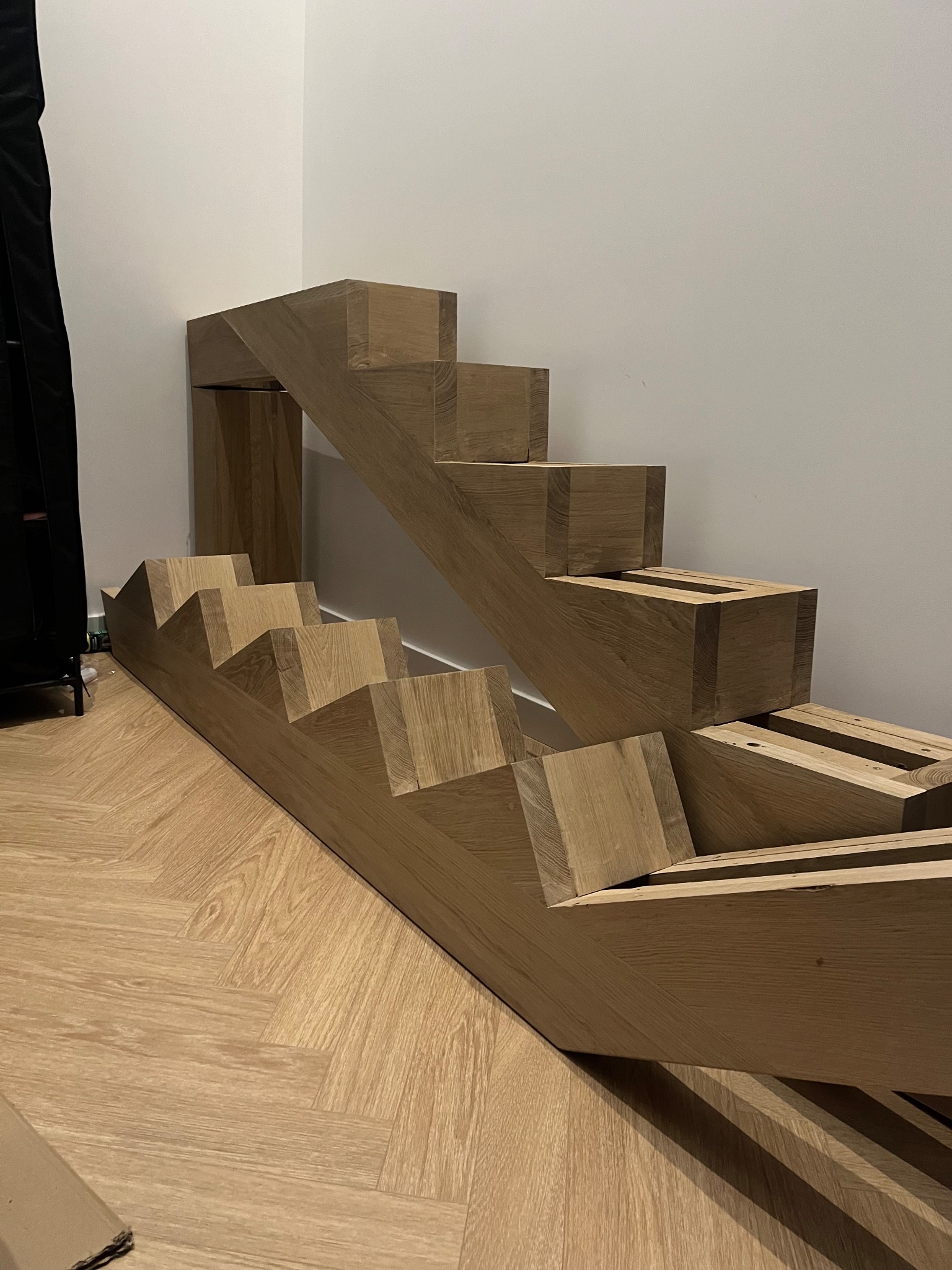 Elementy drewnianych schodów w trakcie montażu, widoczne na podłodze w jodełkę, z fragmentem zamontowanej konstrukcji przy ścianie. Jasne drewno, surowy stan.