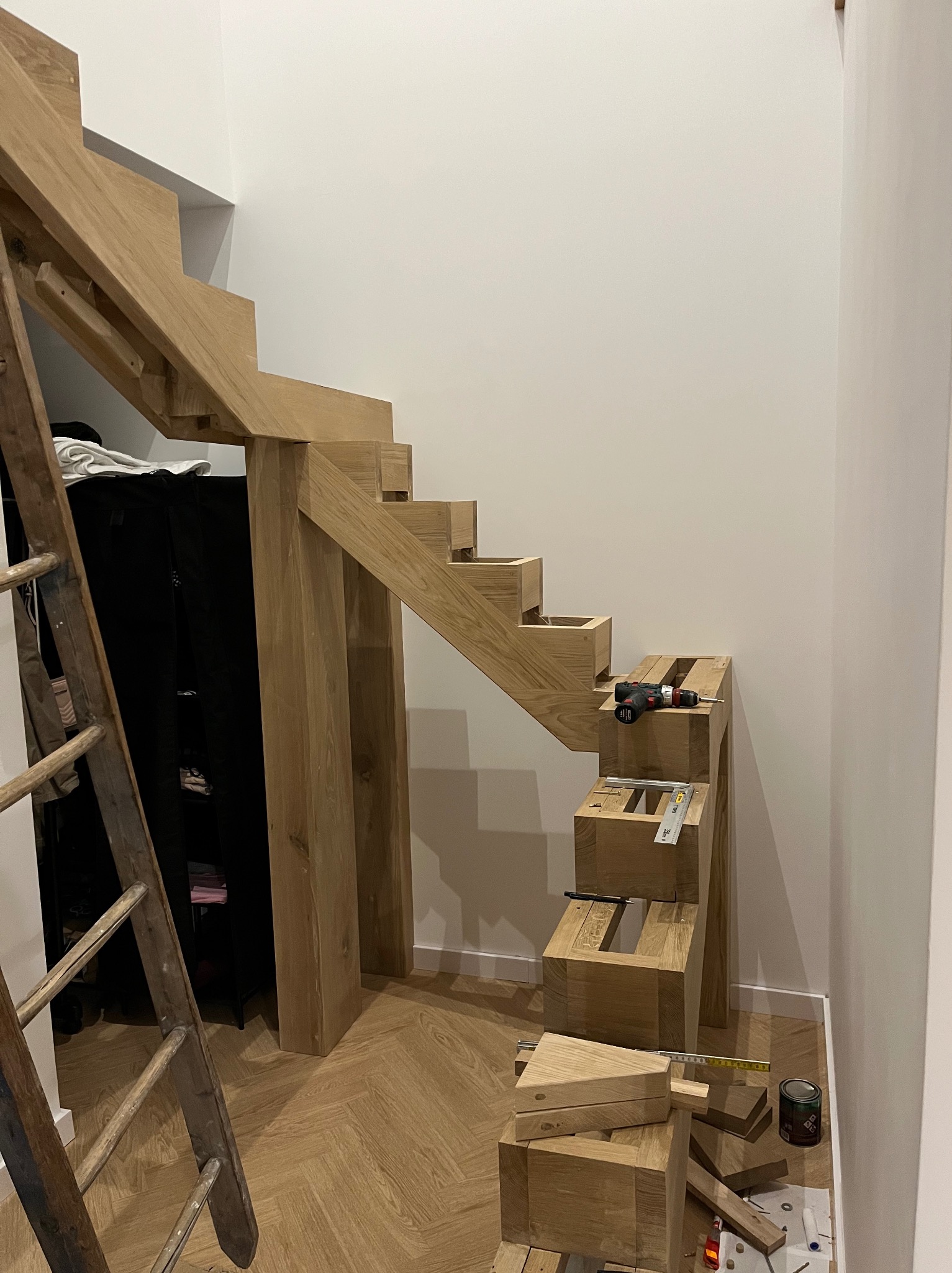 Surowe, dębowe schody w trakcie montażu, z widocznymi narzędziami i elementami konstrukcyjnymi. Stara drabina oparta o ścianę. Parkiet w jodełkę na podłodze.