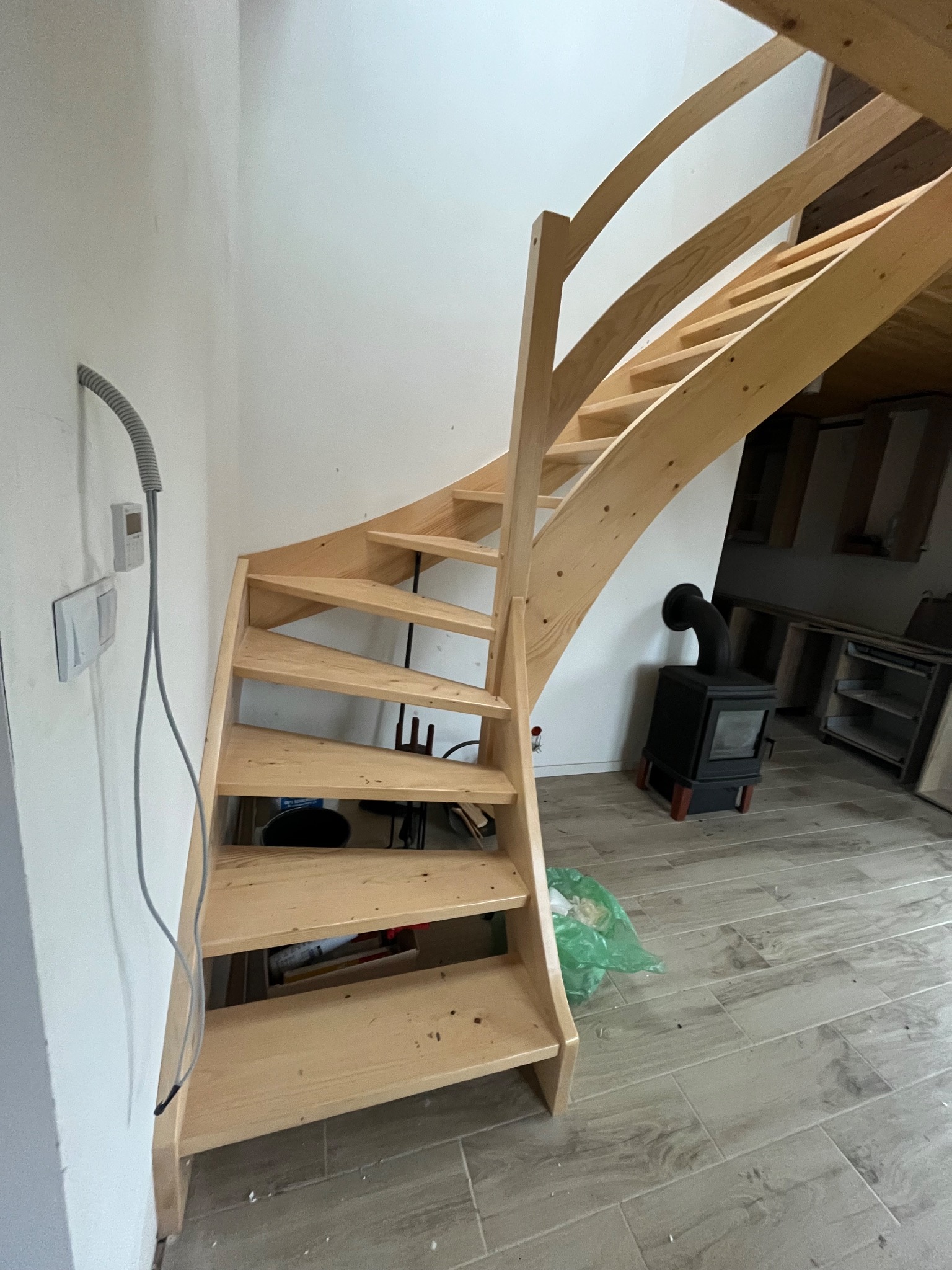 Spiralne schody drewniane z jasnego drewna, z widocznym piecem wolnostojącym w tle. Ujęcie z dołu, ukazujące konstrukcję i detale wykończenia.