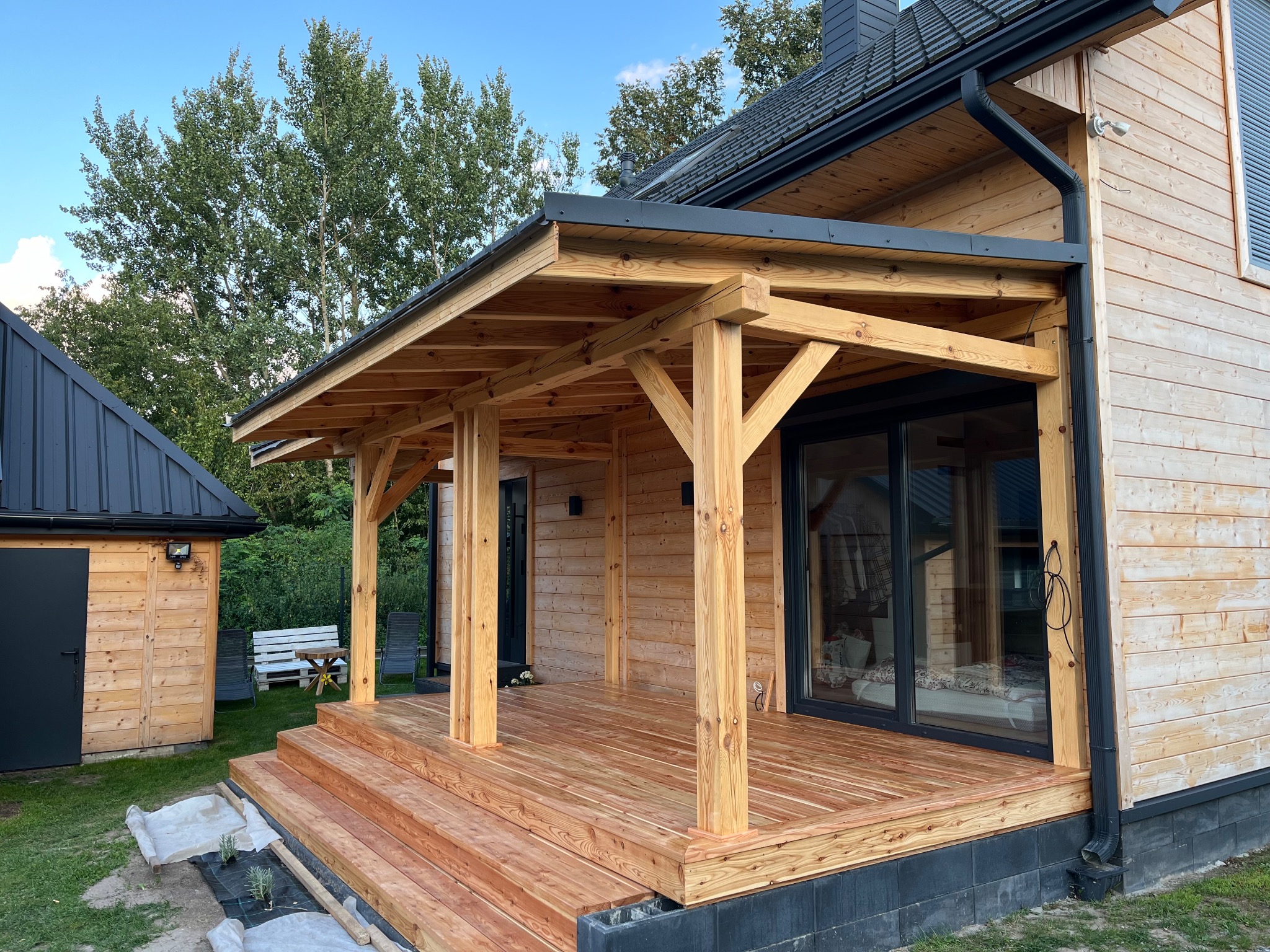 Zadaszony taras z drewna przy domu z drewnianą elewacją. Widoczne schody wejściowe, elementy konstrukcyjne z jasnego drewna i ciemne wykończenia dachu.