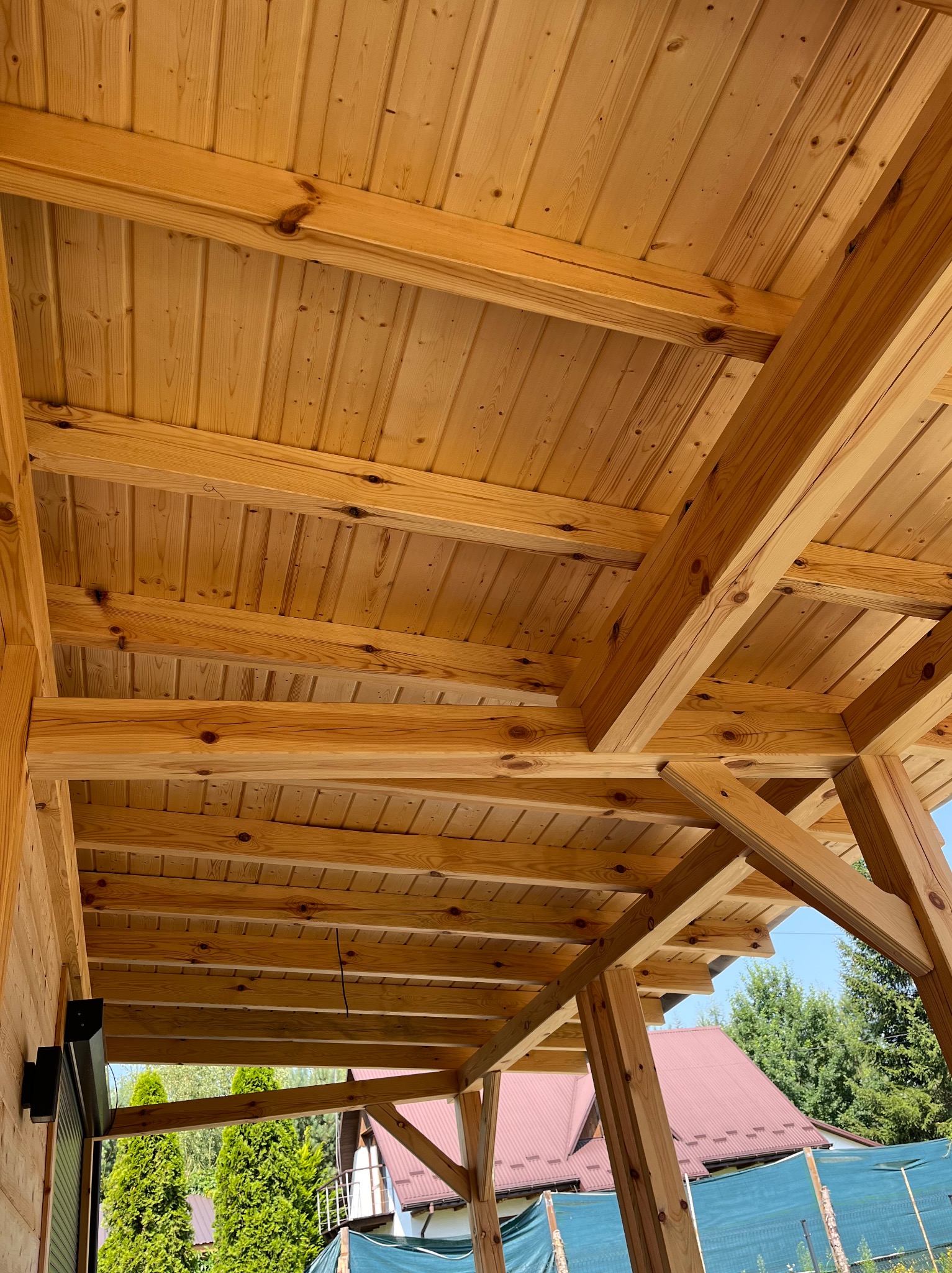 Jasna, drewniana konstrukcja z belek i desek, prawdopodobnie zadaszenie tarasu lub altany, widziana z dołu pod kątem, z fragmentem zieleni w tle.