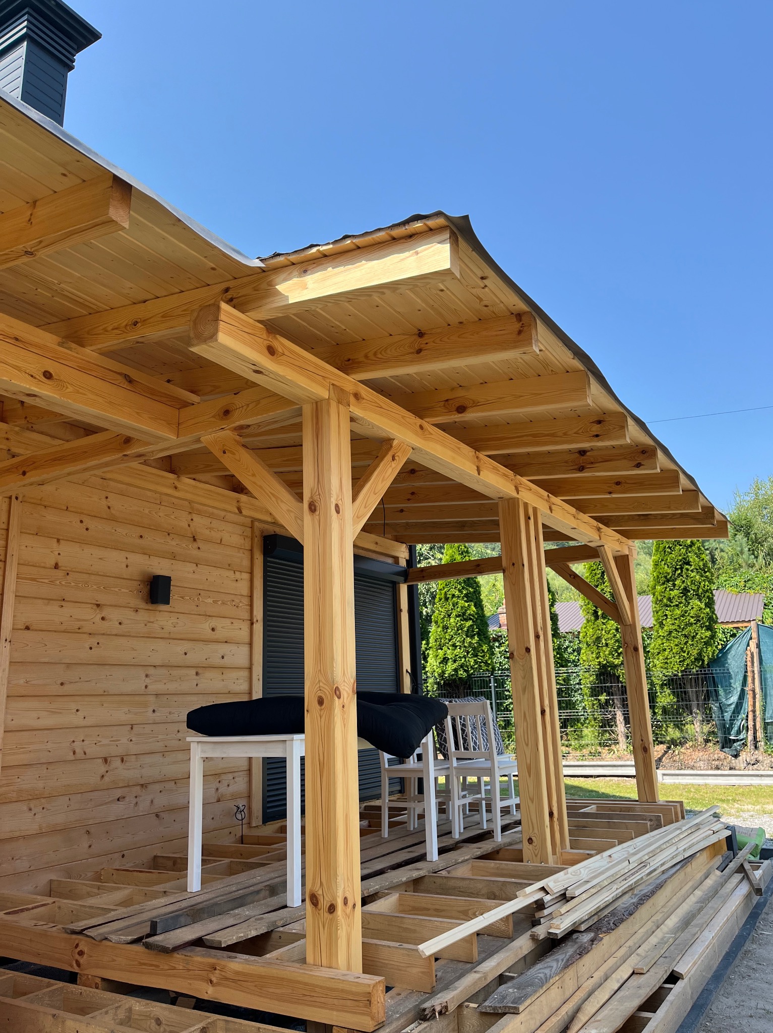 Surowa, drewniana konstrukcja zadaszenia tarasu w trakcie budowy, z widocznymi elementami podłogi i meblami ogrodowymi w tle, na tle błękitnego nieba.