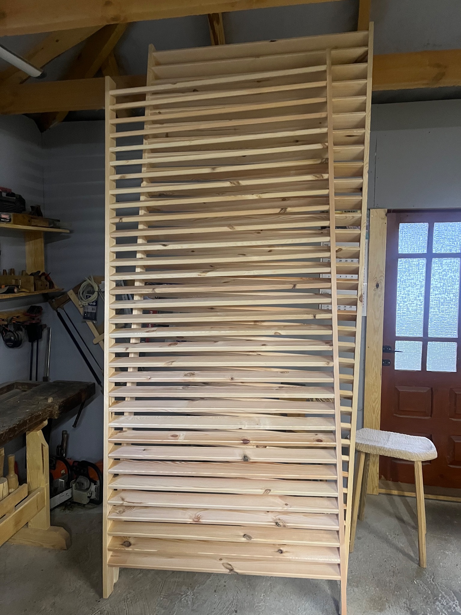 Surowa, drewniana konstrukcja z listew, prawdopodobnie element większej całości, w warsztacie. Widoczne narzędzia i taboret.