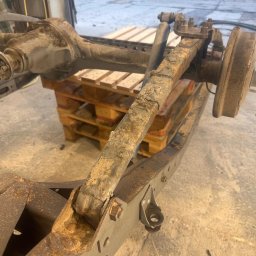 Magnaris - Zardzewiałe podwozie samochodu na palecie w warsztacie. Widoczne ślady korozji i zużycia. Elementy zawieszenia i układu hamulcowego. Przygotowanie do renowacji antykorozyjnej.