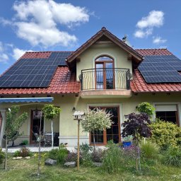 Magnaris - Dom jednorodzinny z panelami fotowoltaicznymi na czerwonym dachu, balkonem i zadbanym ogrodem w słoneczny dzień. Widoczne błękitne niebo z chmurami.