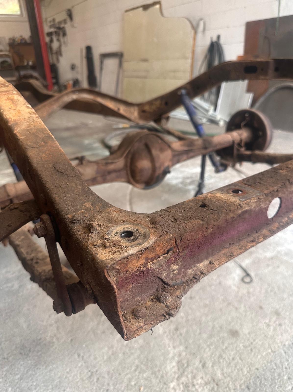 Zardzewiała rama samochodu z widocznymi śladami korozji, ukazana z bliska w warsztacie. Elementy zawieszenia i osi w tle. Widoczne ślady wcześniejszego lakierowania.