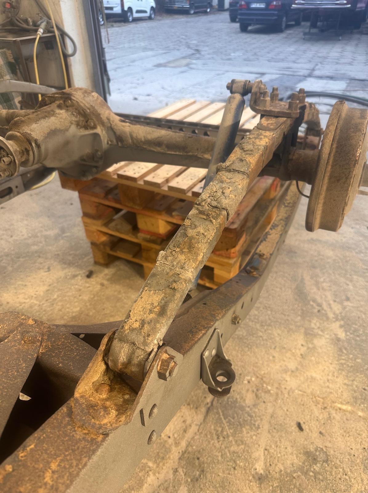 Zardzewiałe podwozie samochodu na palecie w warsztacie. Widoczne ślady korozji i zużycia. Elementy zawieszenia i układu hamulcowego. Przygotowanie do renowacji antykorozyjnej.