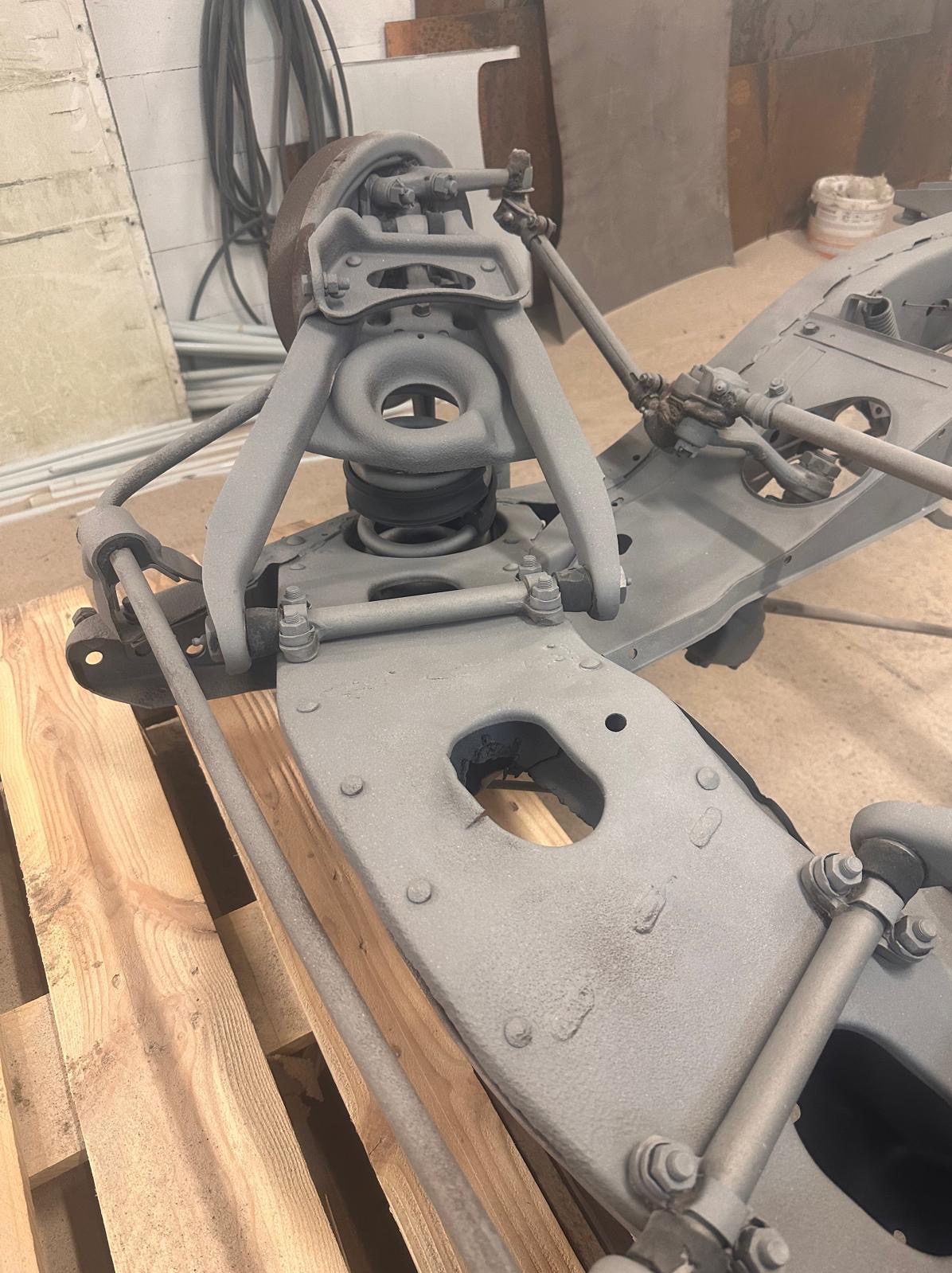 Szare podwozie samochodu po piaskowaniu, umieszczone na drewnianej palecie. Widoczne elementy zawieszenia i układu kierowniczego. Detaliczny widok na odnowioną konstrukcję.