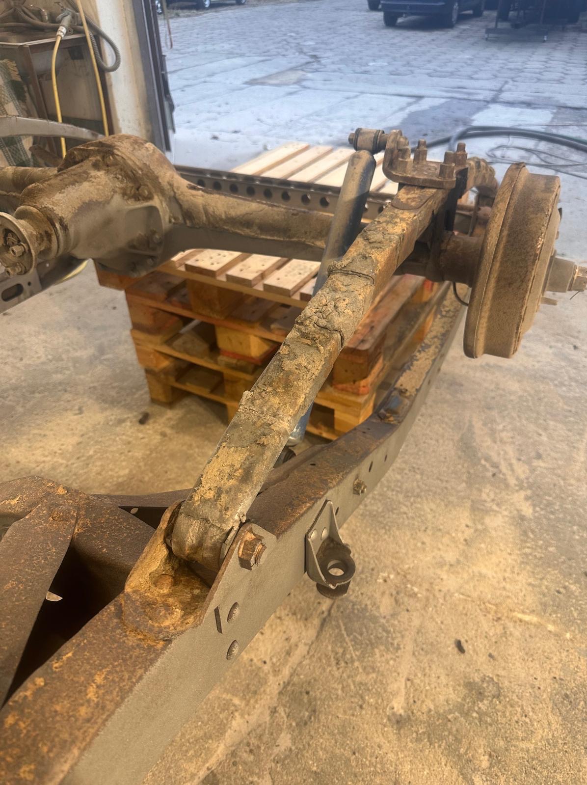 Zardzewiałe podwozie samochodu na palecie w warsztacie, widoczne elementy zawieszenia i mocowania. Detaliczny widok na korozję i zużycie.