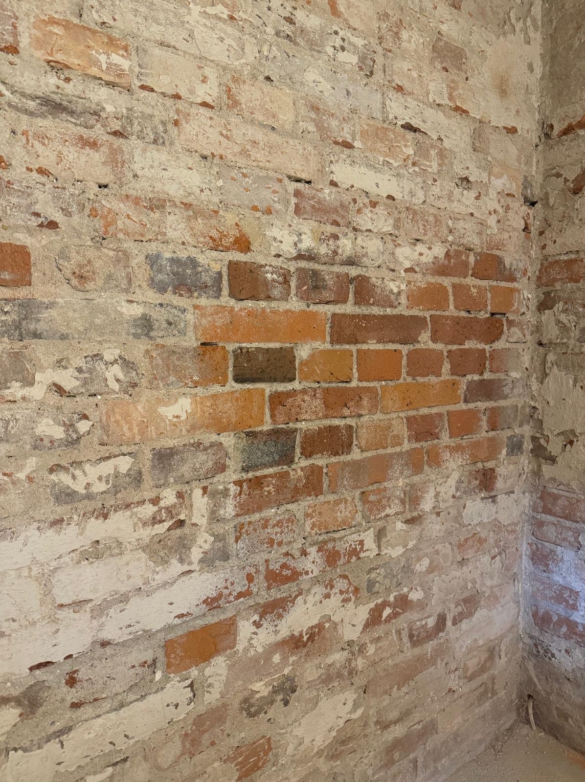 Ceglana ściana z widocznymi śladami starej farby i zaprawy, ukazująca surowy, industrialny charakter wnętrza. Fragment narożnika podkreśla trójwymiarowość tekstury.