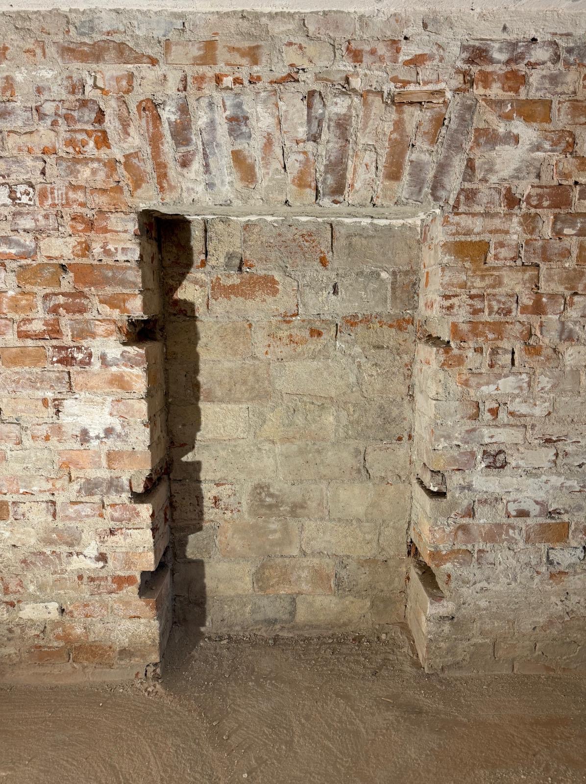 Niezatynkowana ściana z czerwonej cegły z wnęką z łukowym nadprożem, fragment podłogi. Widoczne ślady napraw i uzupełnień zaprawą. Surowy, industrialny charakter.