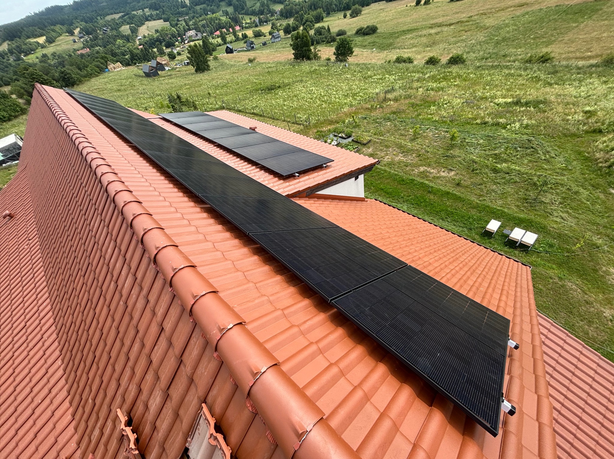 Panele fotowoltaiczne na dachu z czerwonej dachówki, widok z góry na zielone wzgórza w tle. Instalacja solarna na tle krajobrazu.