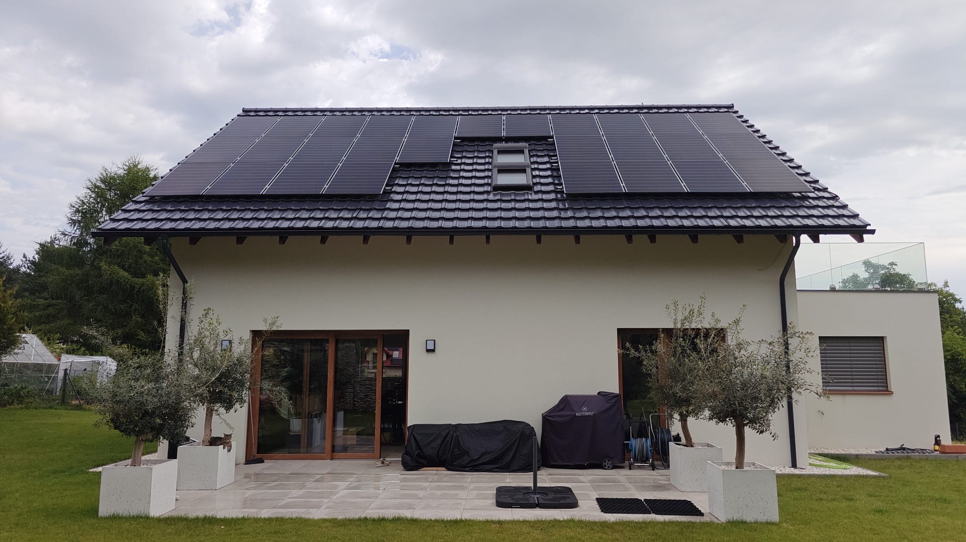 Dom jednorodzinny z panelami fotowoltaicznymi na dachu, widok z ogrodu. Na pierwszym planie drzewka w donicach, w tle zielona trawa i drzewa.