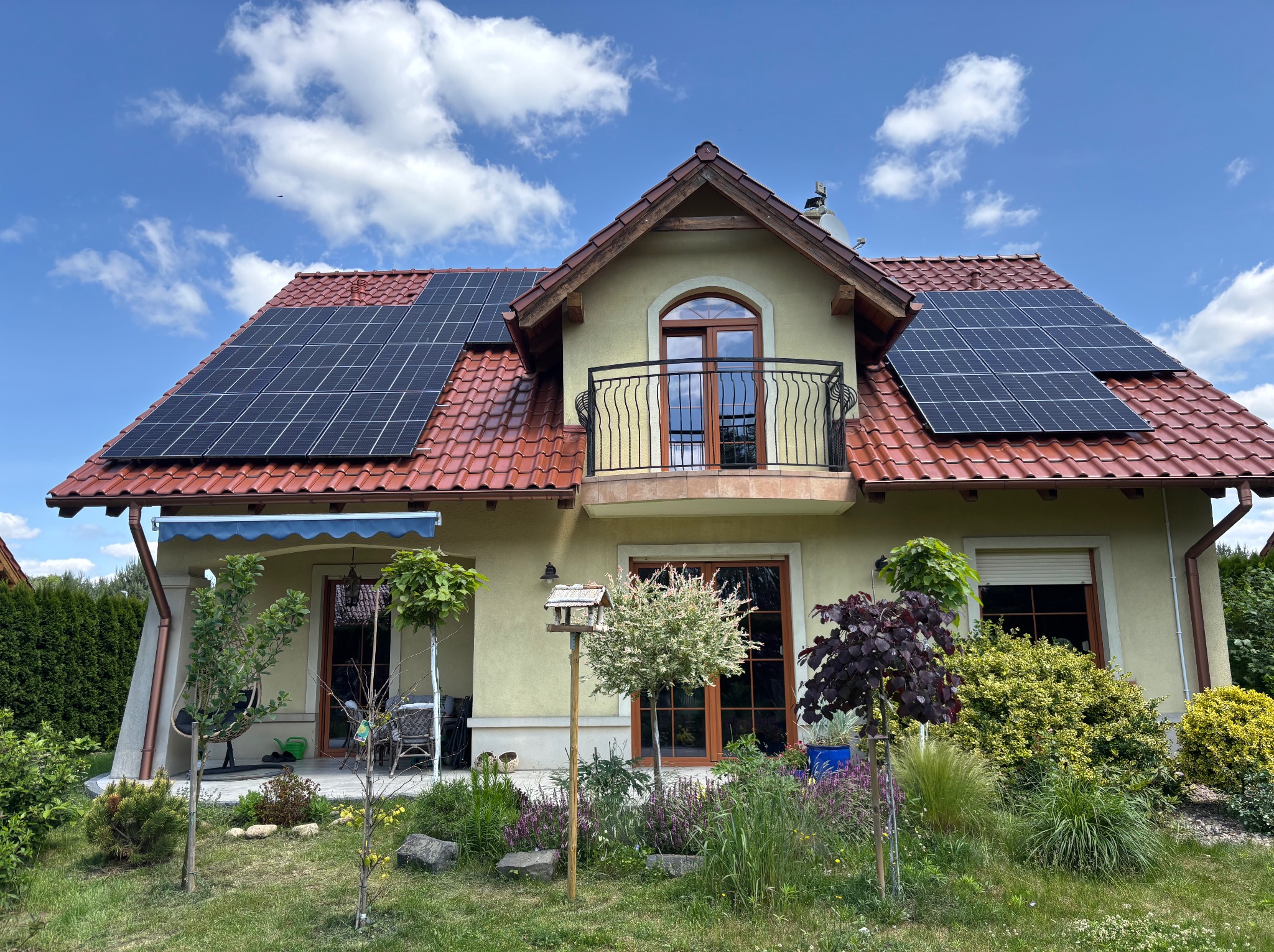 Dom jednorodzinny z panelami fotowoltaicznymi na czerwonym dachu, balkonem i zadbanym ogrodem w słoneczny dzień. Widoczne błękitne niebo z chmurami.