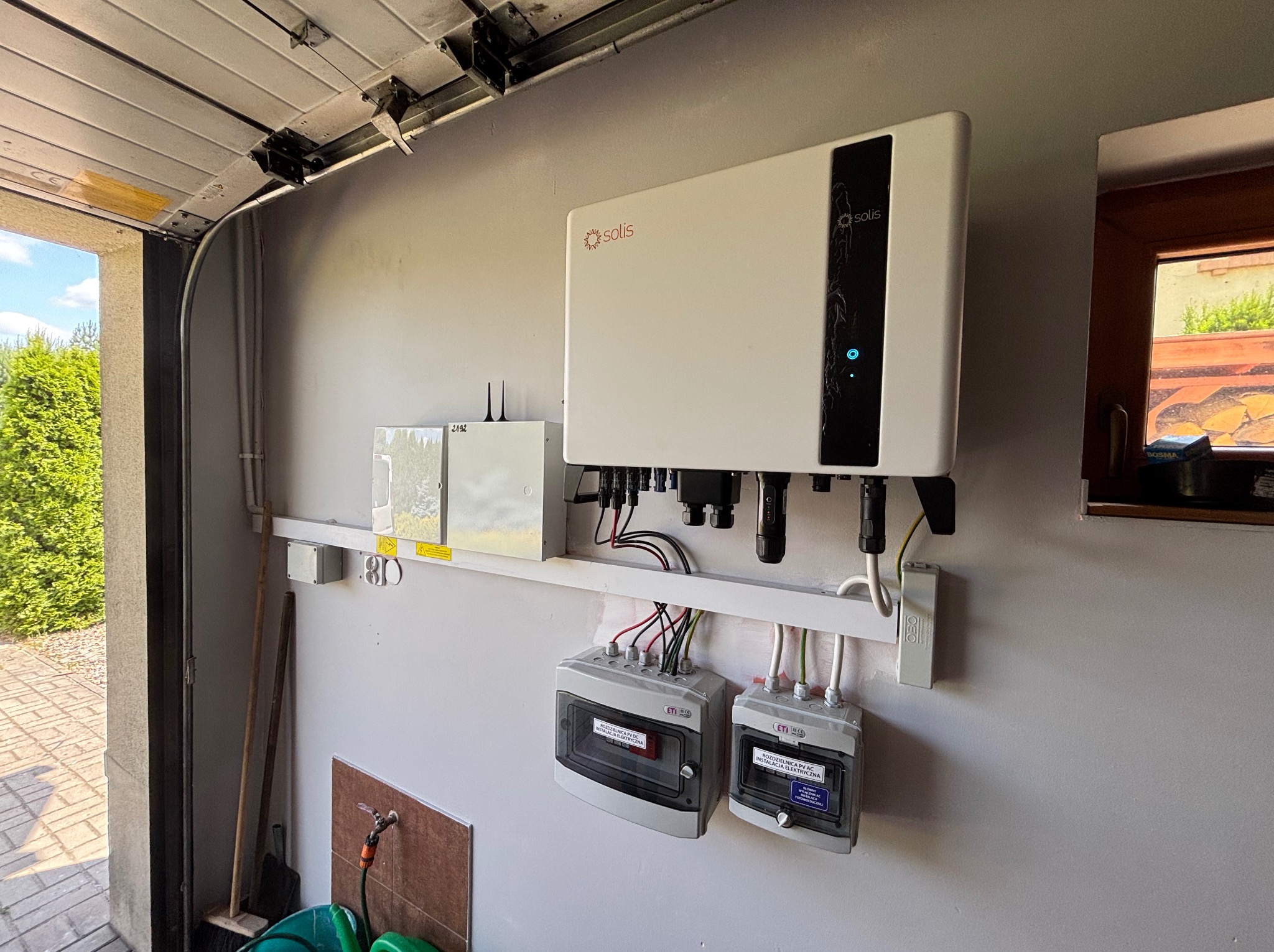 Biały inwerter Solis zamontowany na ścianie garażu, z widocznymi rozdzielnicami PV AC i okablowaniem. Elementy instalacji fotowoltaicznej wewnątrz budynku.