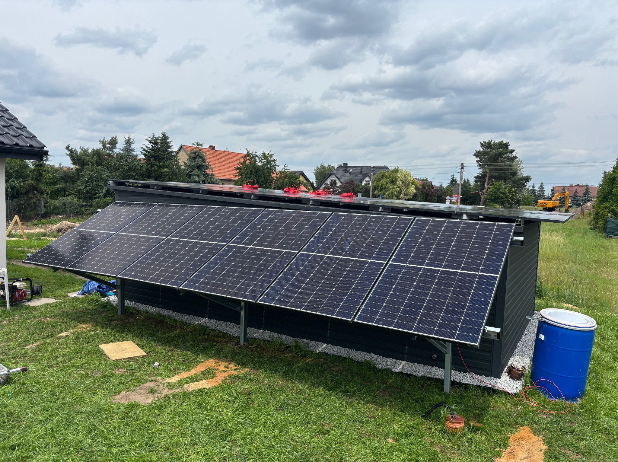 Gotowa instalacja fotowoltaiczna na dachu budynku gospodarczego, widok z poziomu gruntu, w tle domy jednorodzinne i pochmurne niebo.