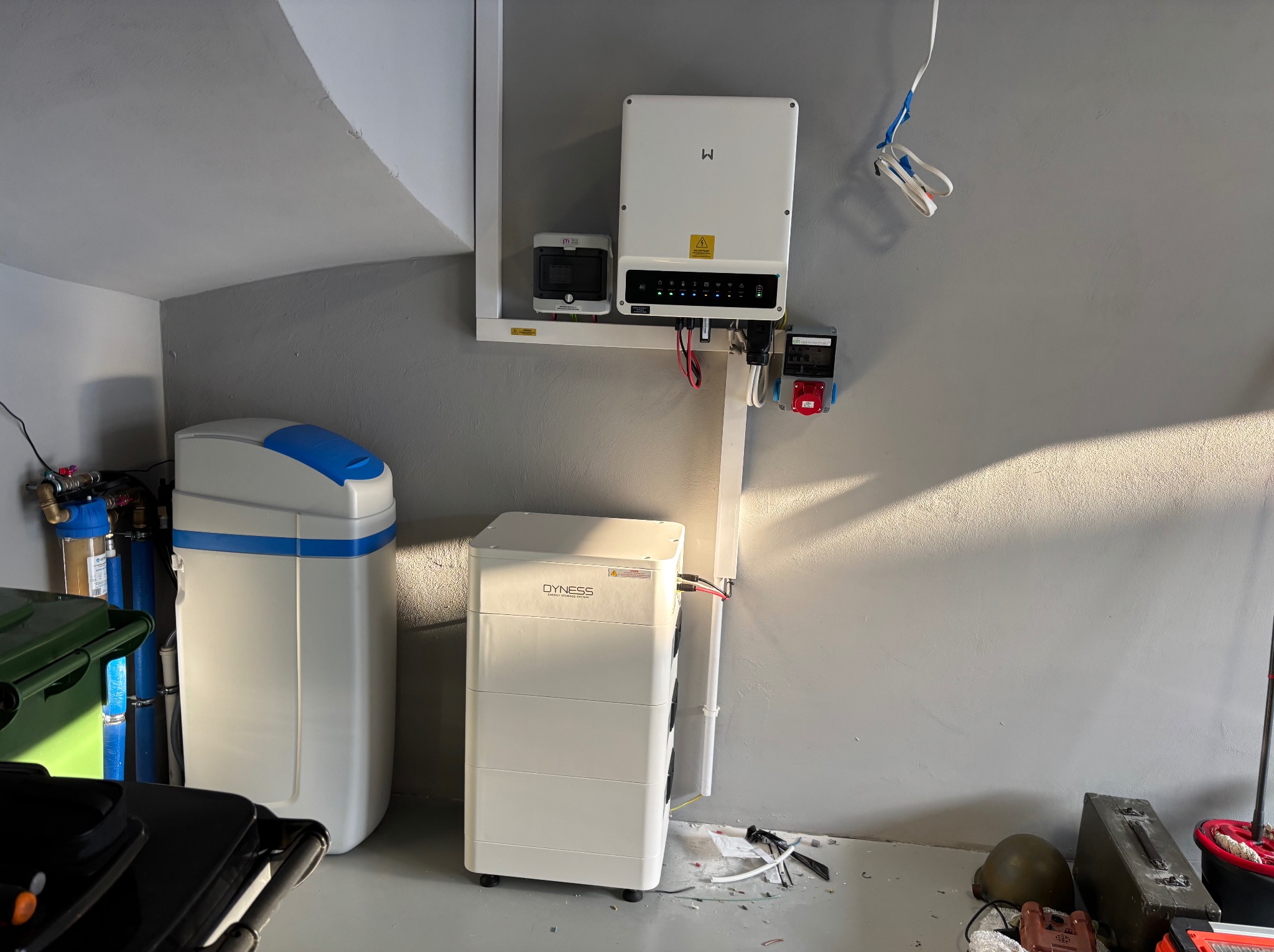 Kompletna instalacja fotowoltaiczna w garażu: inwerter, licznik, akumulator Dyness, filtr wody. Widoczne przewody i narzędzia, co sugeruje etap końcowy montażu.