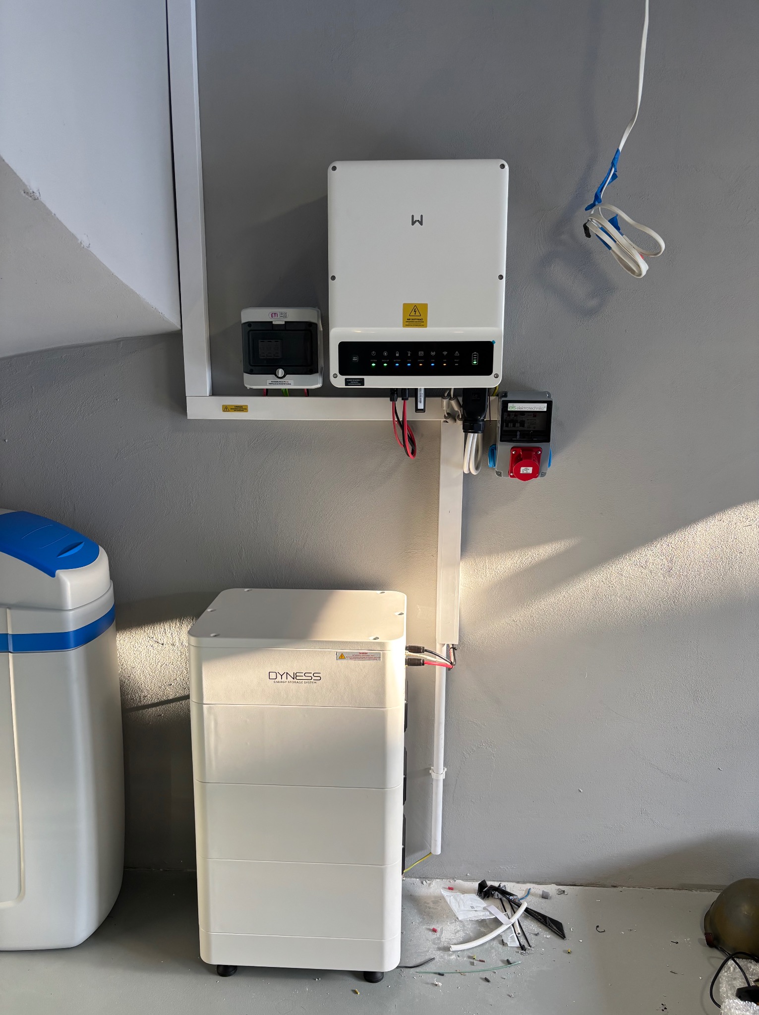 Zainstalowany system fotowoltaiczny w pomieszczeniu: biały inwerter, akumulator Dyness, licznik energii i gniazdo siłowe na szarej ścianie. Widoczne przewody i elementy instalacji.