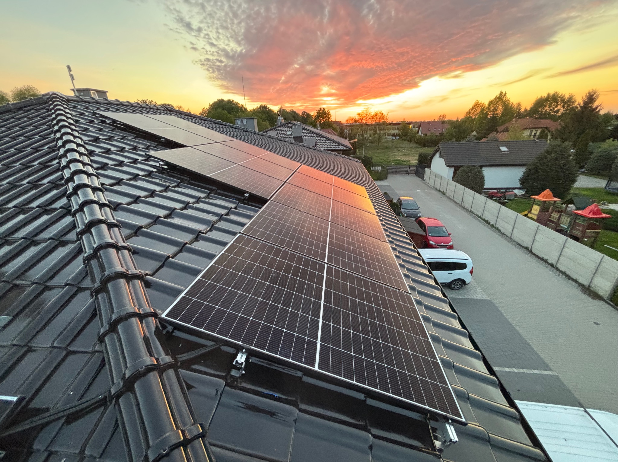 Panele fotowoltaiczne na dachu domu, widok z góry, zachód słońca z pomarańczowymi chmurami w tle. Widoczne samochody i fragment ulicy.