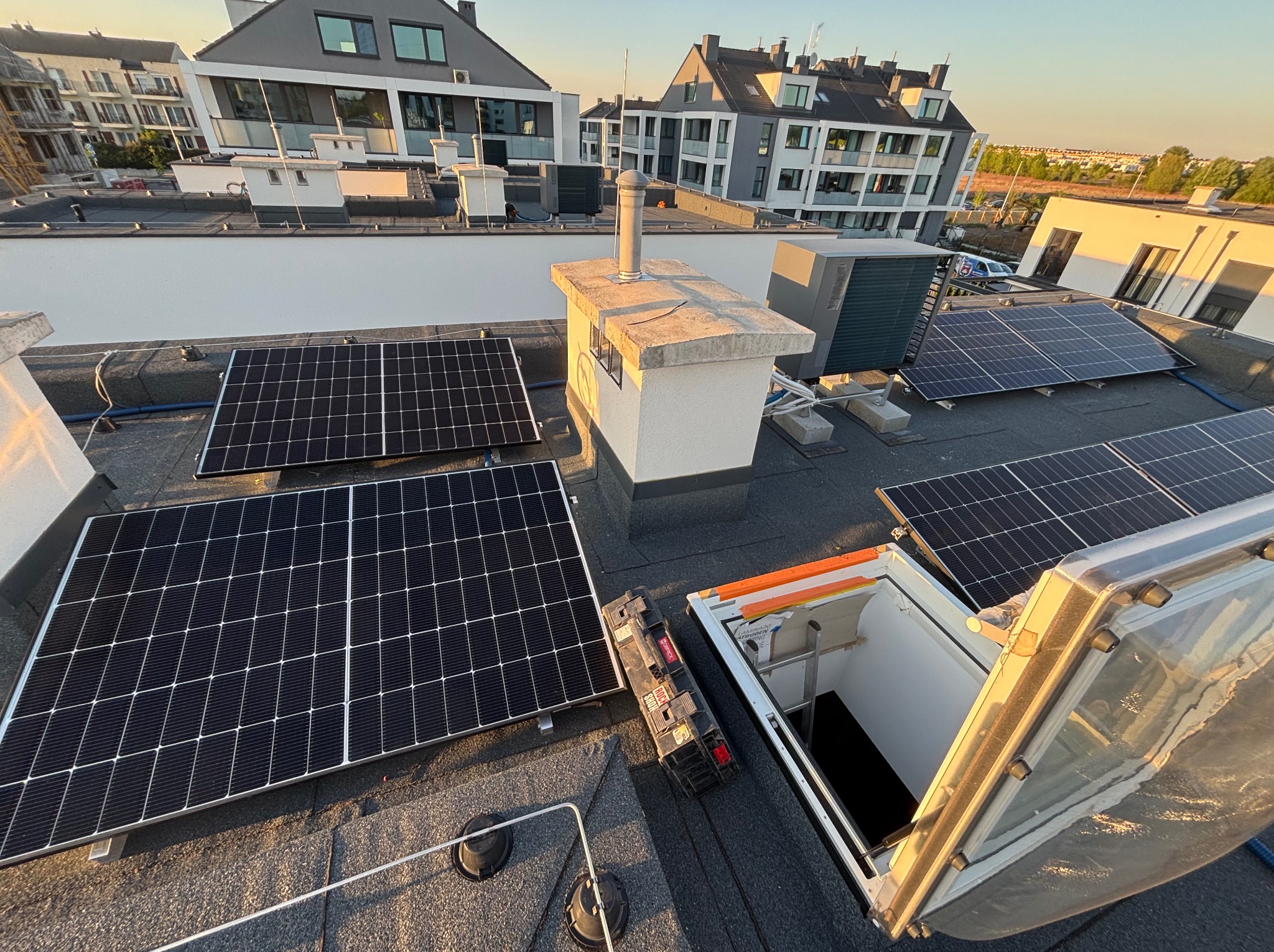 Instalacja paneli fotowoltaicznych na dachu budynku wielorodzinnego, widok z góry na panele, komin wentylacyjny i otwarty właz dachowy z drabiną.