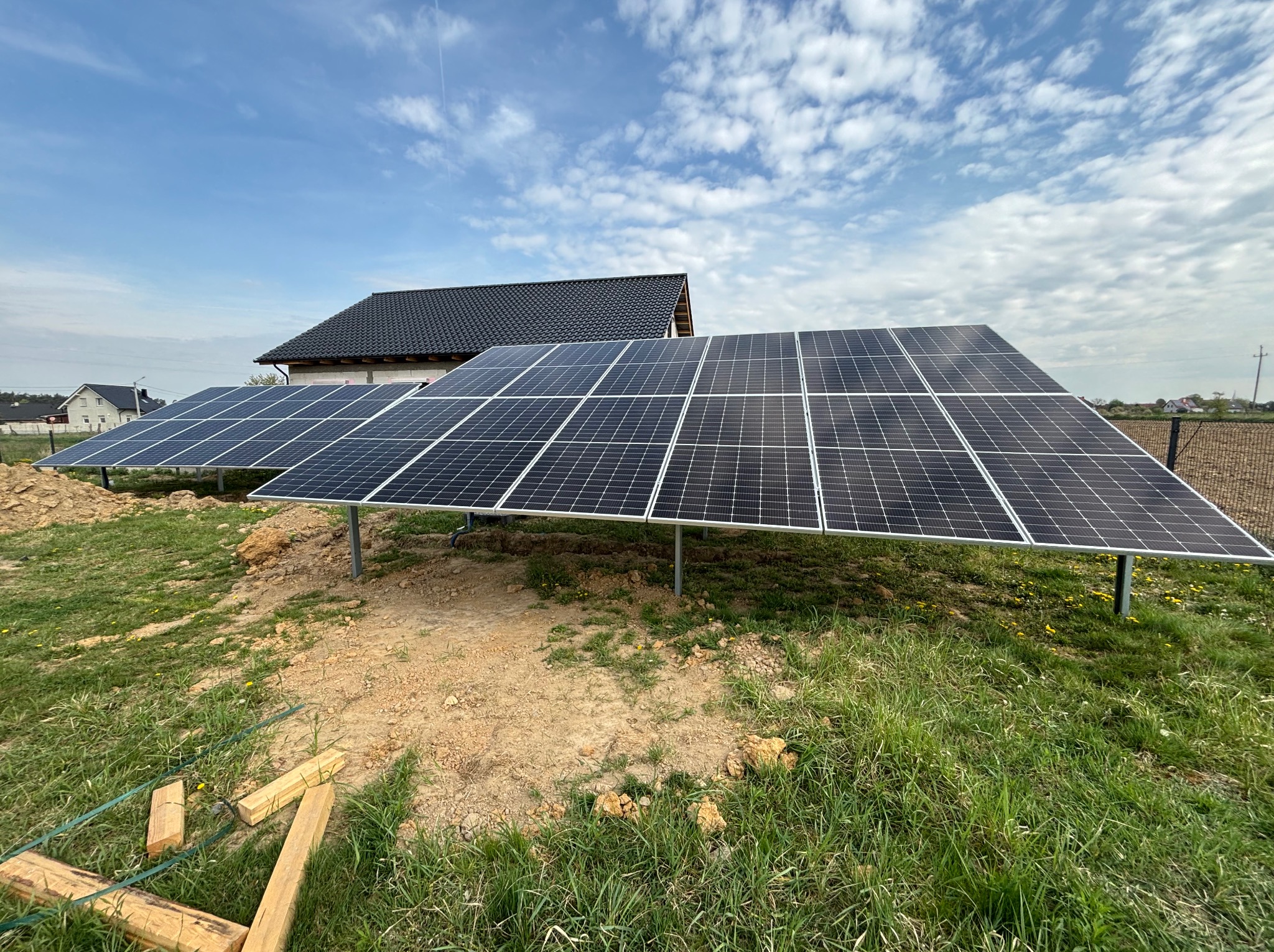 Instalacja fotowoltaiczna na gruncie, panele ustawione pod kątem, w tle dom z ciemną dachówką i błękitne niebo z chmurami. Widoczne drewniane elementy montażowe.