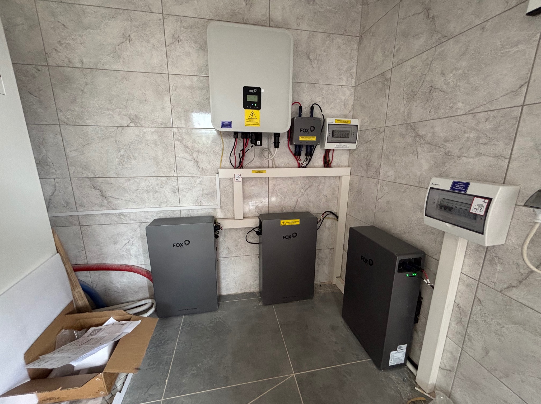 Zainstalowany system fotowoltaiczny FOX: inwerter, akumulatory i rozdzielnica na ścianie z szarych płytek. Widoczne kable i elementy montażowe.