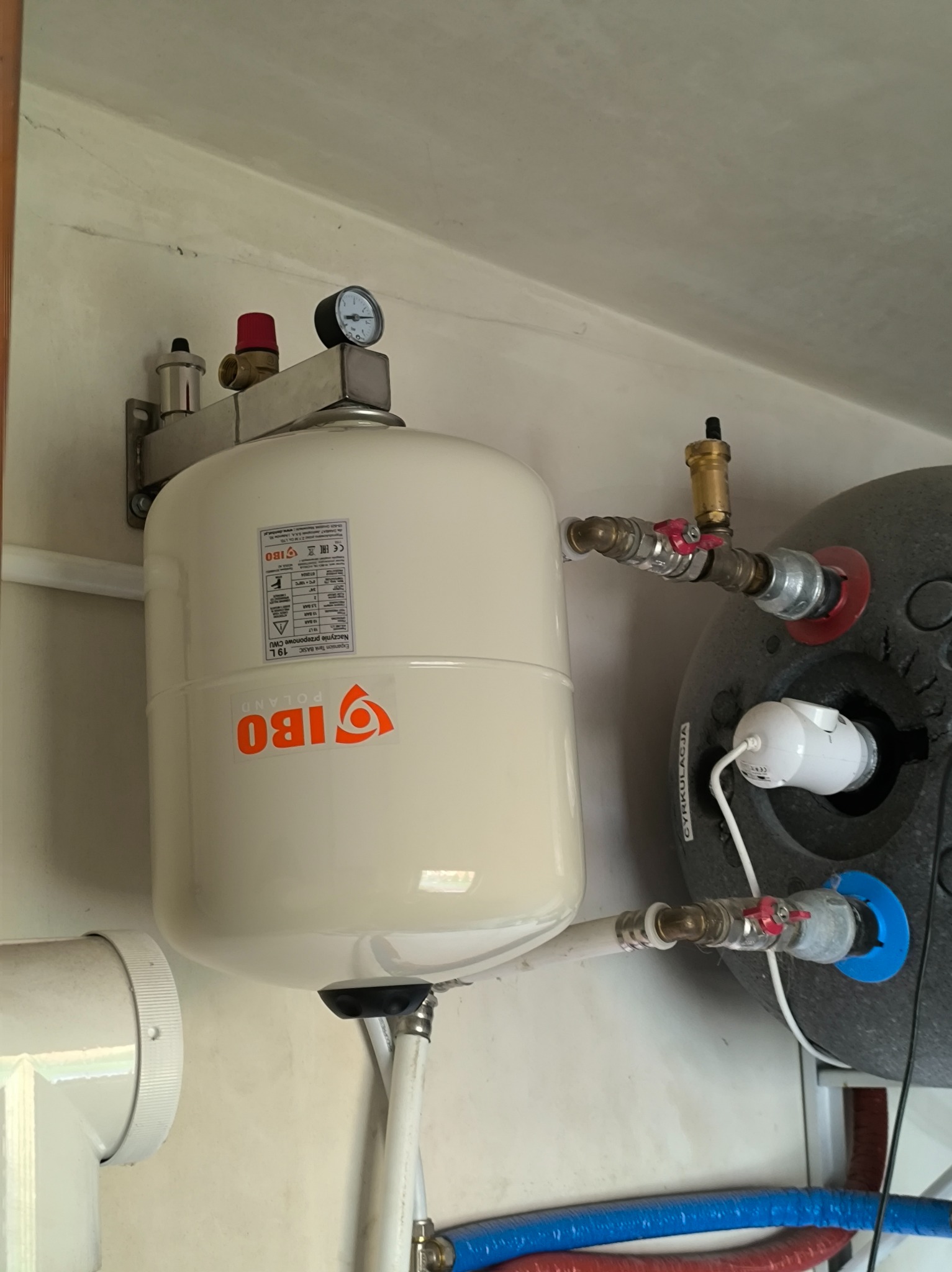 Zamknięte naczynie przeponowe IBO o pojemności 19L, zamontowane na ścianie, z widocznymi przyłączami hydraulicznymi i manometrem. Obok zbiornik akumulacyjny.