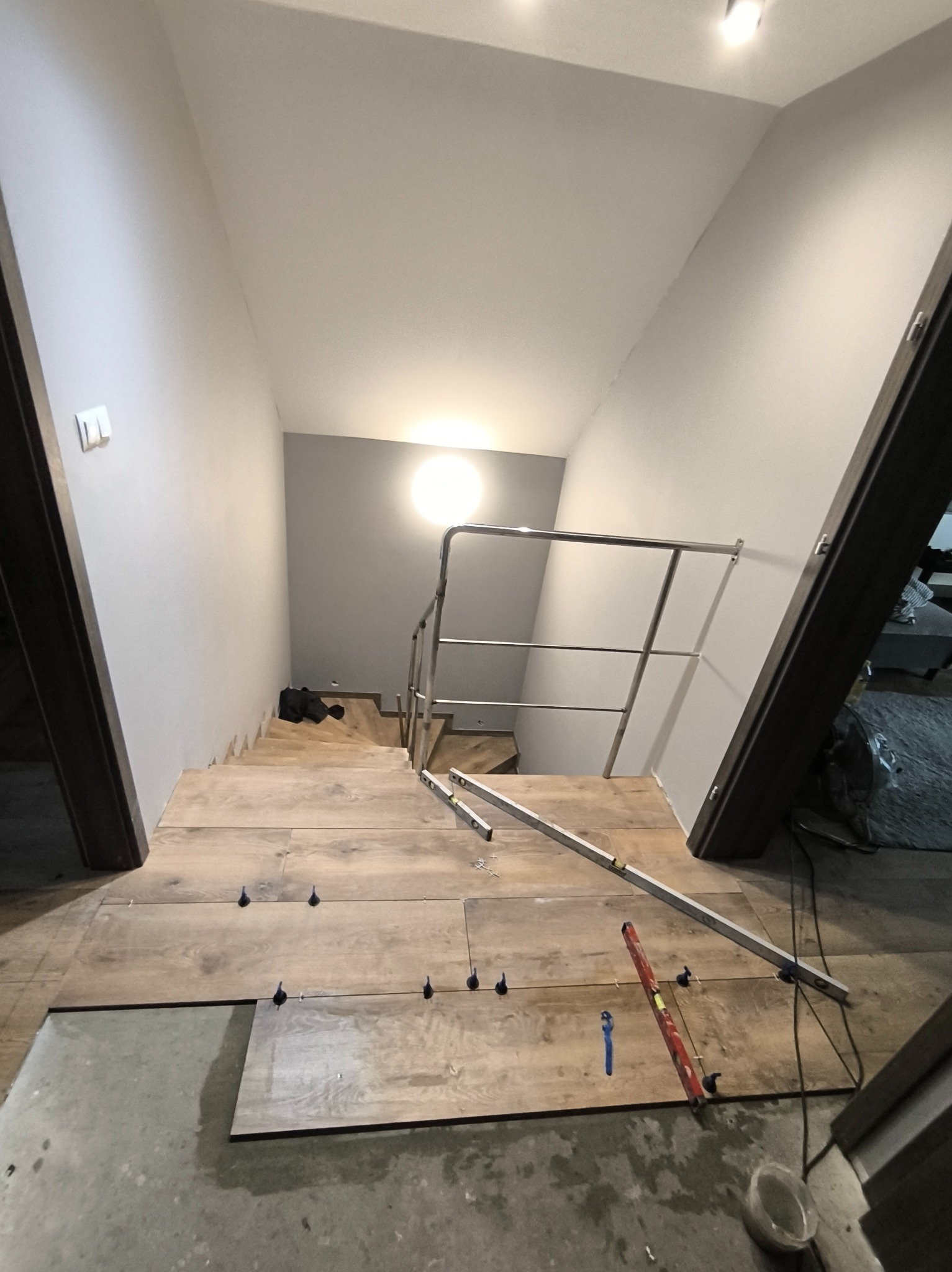 Montaż drewnopodobnych paneli na schodach z metalową barierką. Widoczne poziomice i elementy mocujące. W tle szare ściany i oświetlenie.