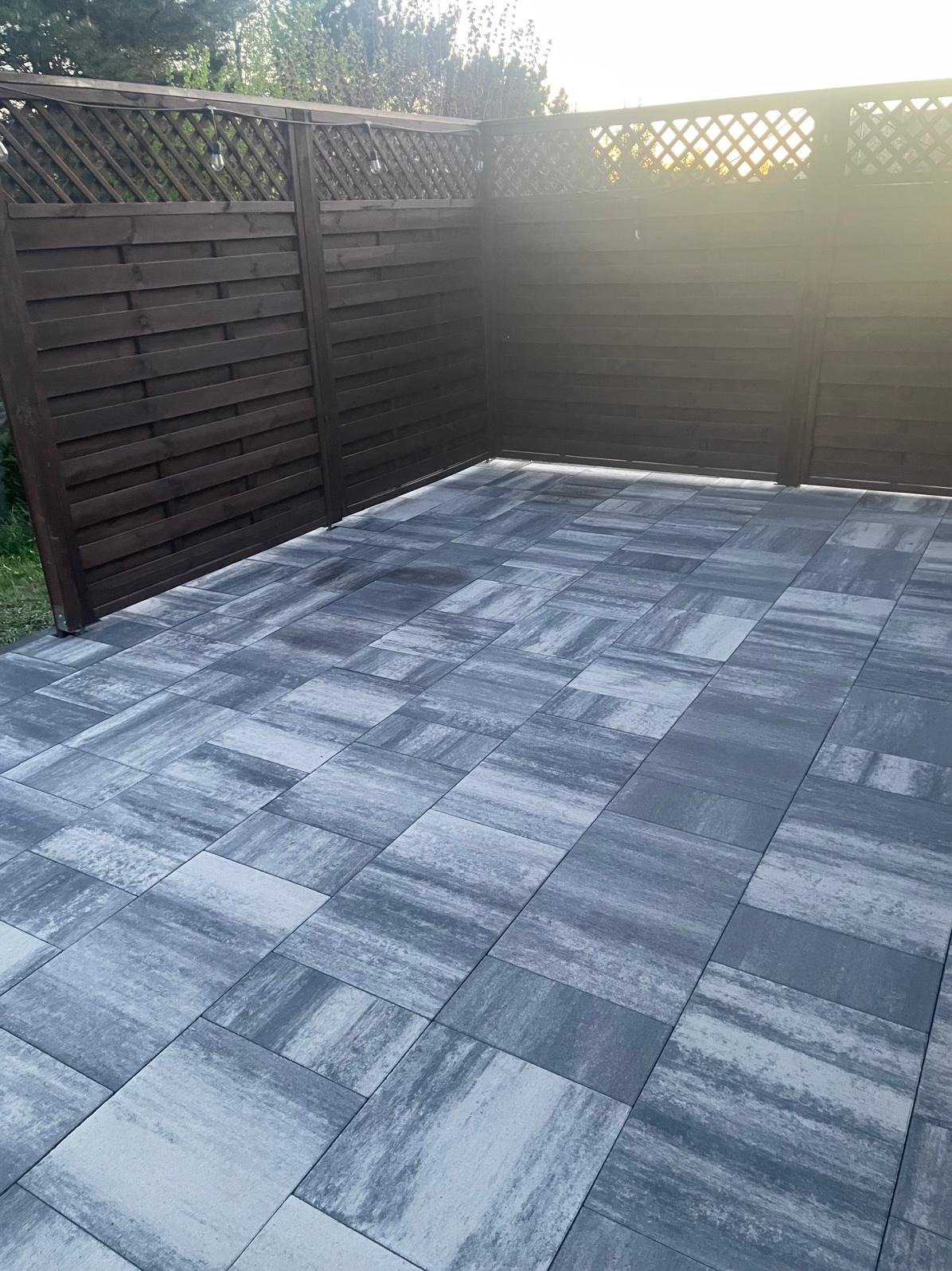 Ułożona szara kostka brukowa na tarasie, otoczona ciemnobrązowym, drewnianym ogrodzeniem z kratką. Słoneczne oświetlenie podkreśla fakturę materiałów.