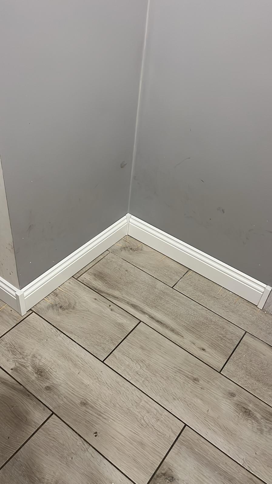 Szare ściany i podłoga z płytek imitujących drewno, wykończone białą listwą przypodłogową w rogu pomieszczenia. Widoczne drobne zabrudzenia na ścianie.