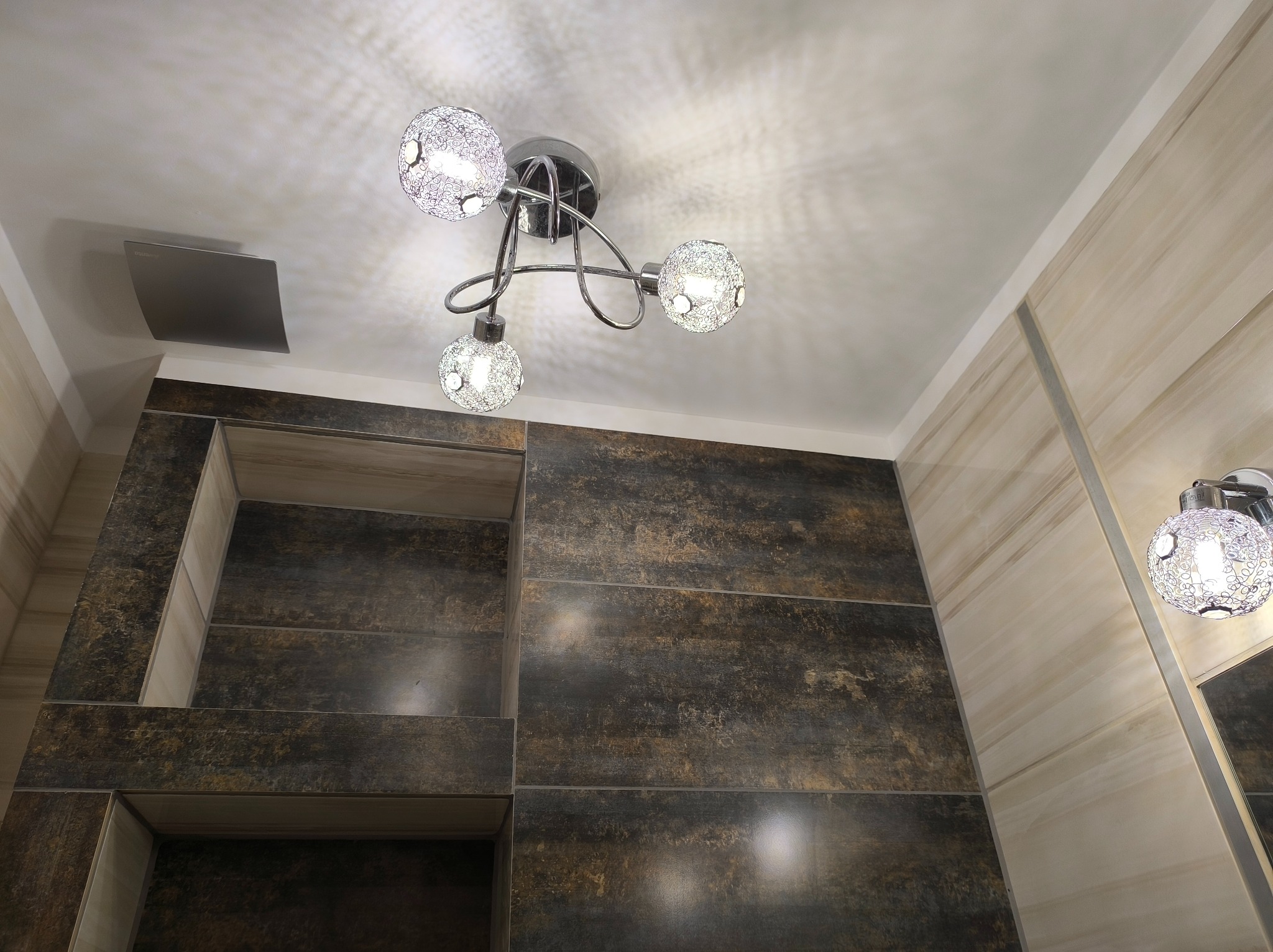 Wykończona łazienka z nowoczesną lampą sufitową i ścienną. Ciemne płytki imitujące drewno z wnękami, jasne płytki na sąsiedniej ścianie. Widok od dołu.