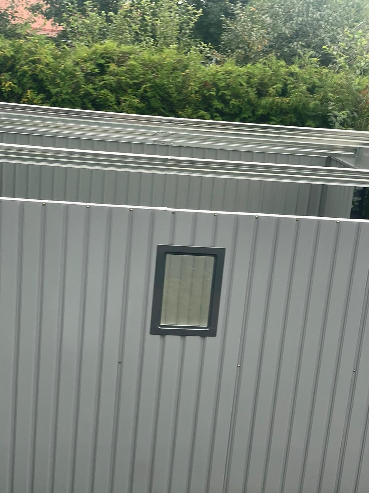 Szara, metalowa konstrukcja z małym oknem w tle zieleni. Widoczne elementy szkieletu i poszycia, sugerujące etap montażu lub budowy.